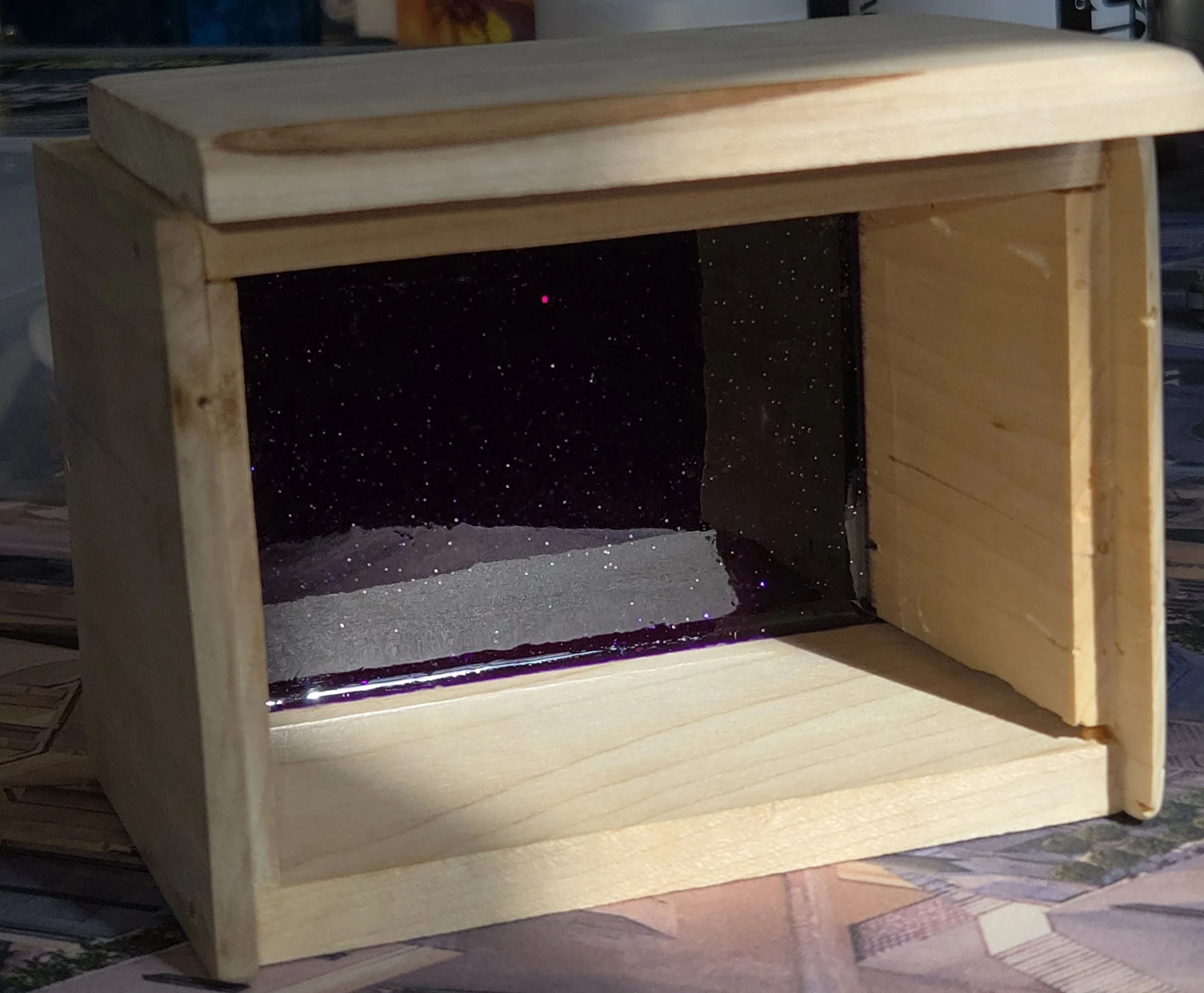 A wooden display case with a glass front, containing a miniature landscape with sand and a starry night sky backdrop.