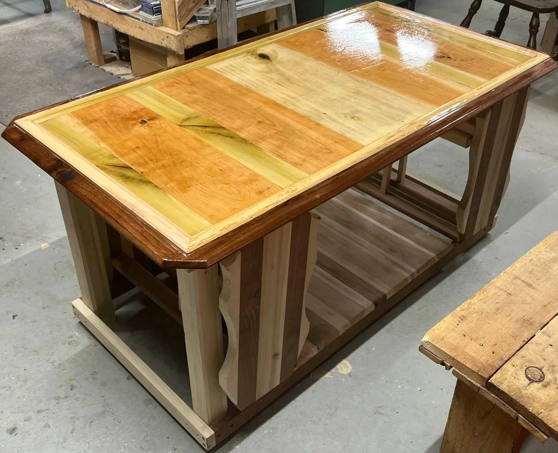 A wooden table with a polished, multicolored wood top and open lower shelf, situated in a workshop.