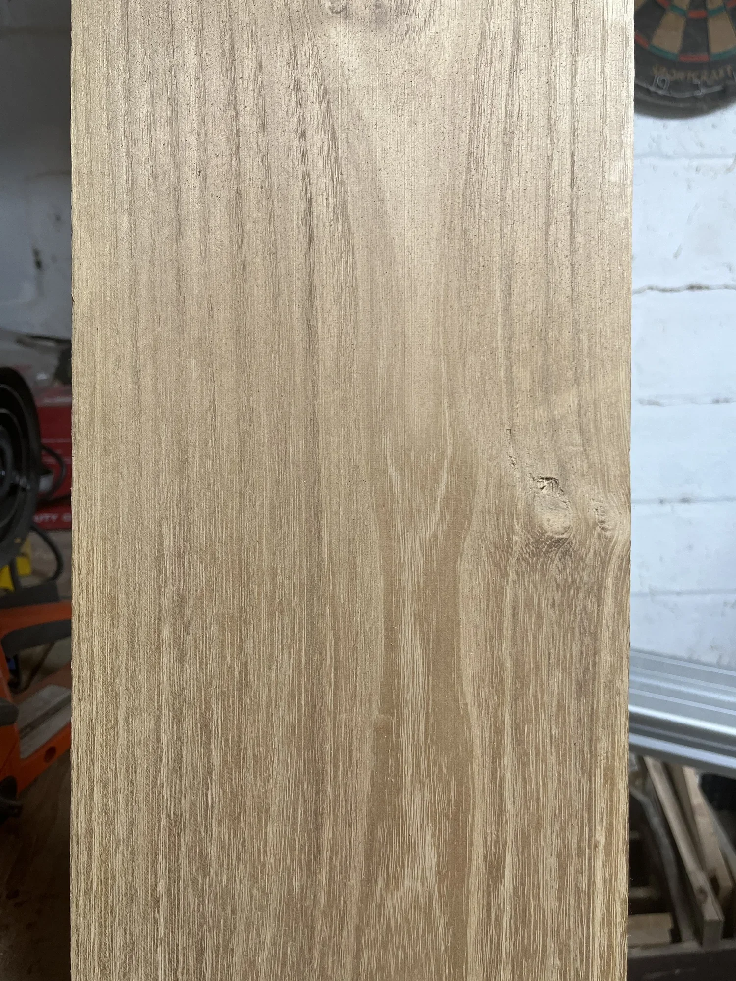 A close-up view of a light-colored wooden board with a visible wood grain pattern and a small knot. Part of a workshop or garage setting with tools and equipment slightly visible in the background.