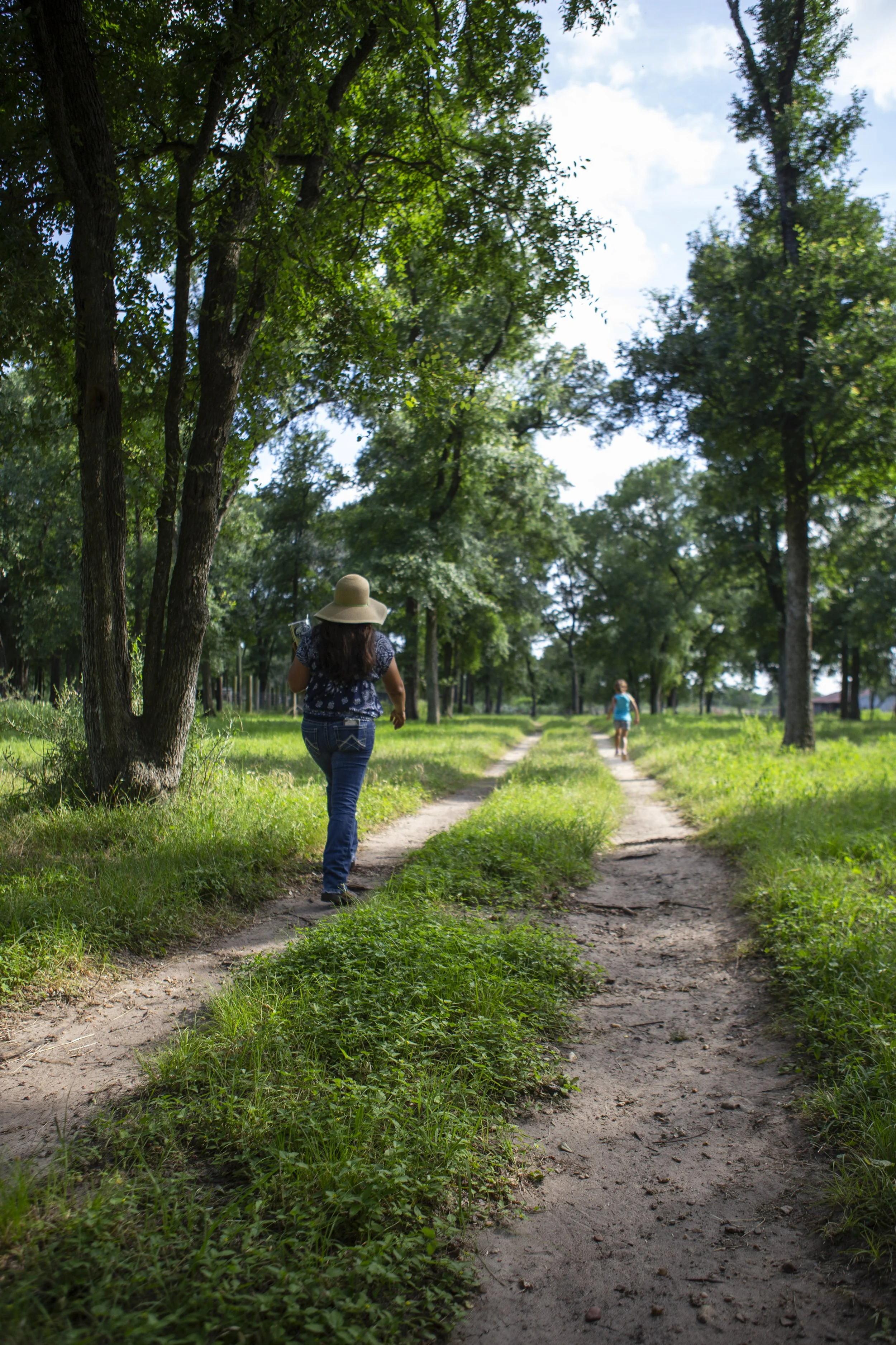 Two women walk along a dirt path in a lush green park with tall trees and a partly cloudy sky.