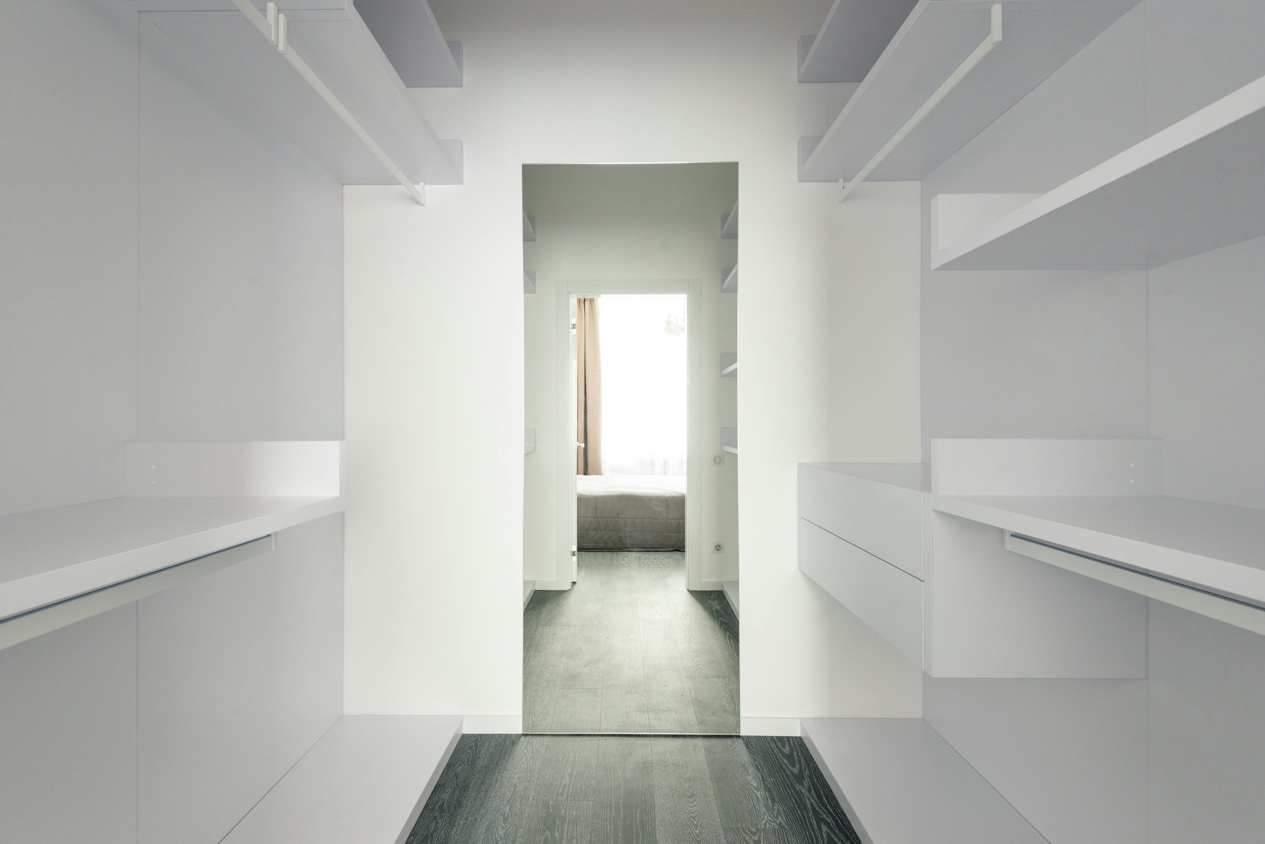 A minimalist white walk-in closet with open shelves on both sides and a mirror at the end reflecting a bedroom with a bed and window.