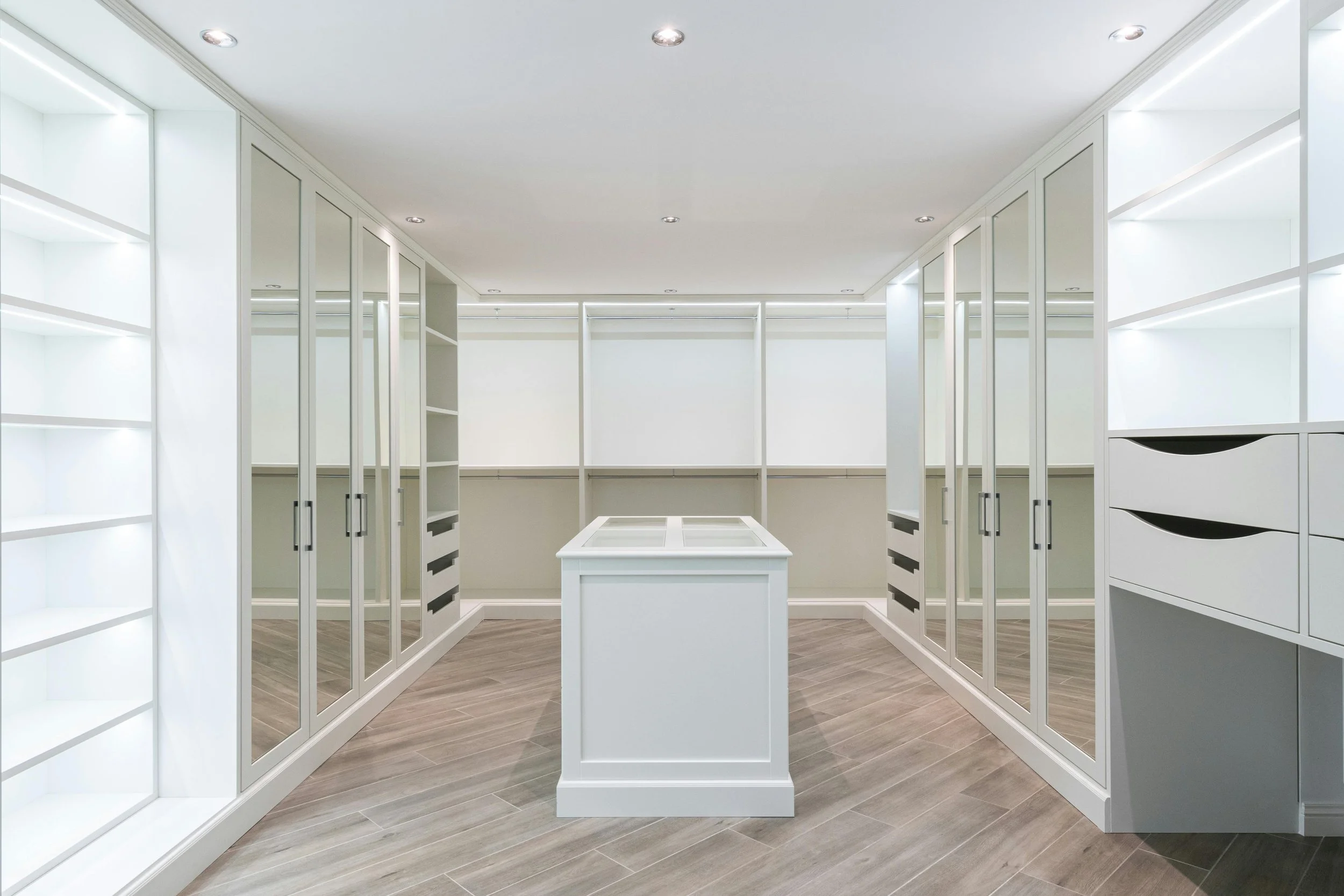 Empty walk-in closet with white shelving, mirrored doors, and a central display table.