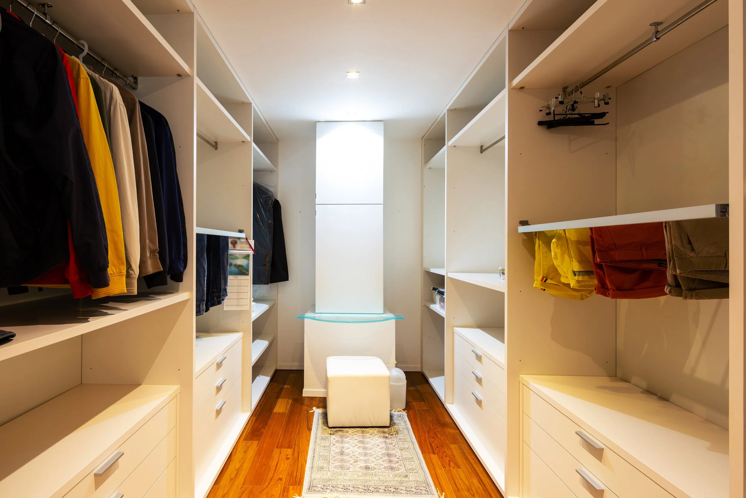 Walk-in closet with white shelves and drawers, displaying clothing such as jackets, pants, and folded garments, with a small rug on a wooden floor and a window at the end.