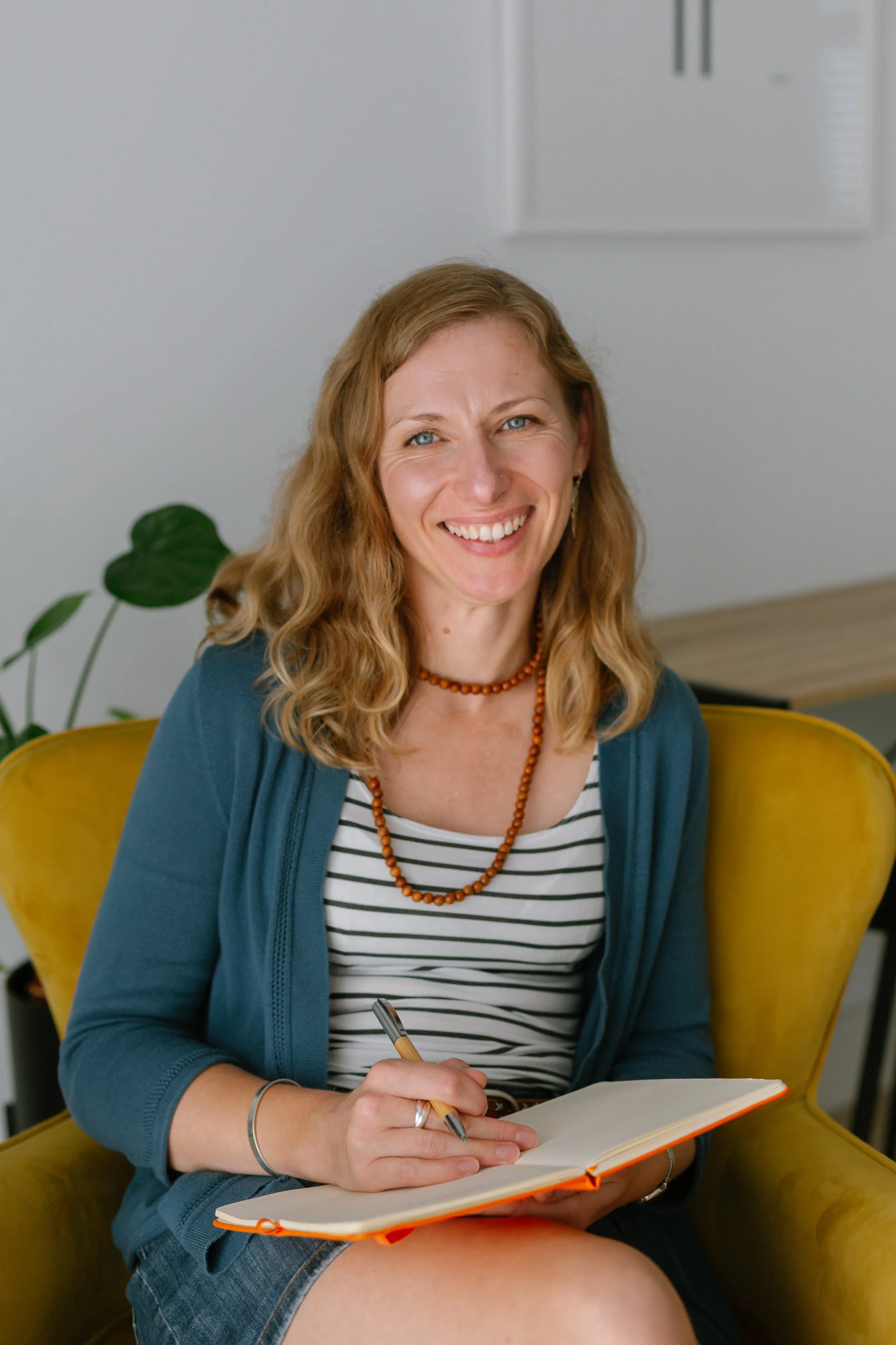 A woman sitting in a yellow chair, smiling, holding a pen, and writing in a notebook. She has wavy blonde hair, blue eyes, wears a striped shirt, a beaded necklace, a blue cardigan, and denim skirt, with a plant and a white wall in the background.
