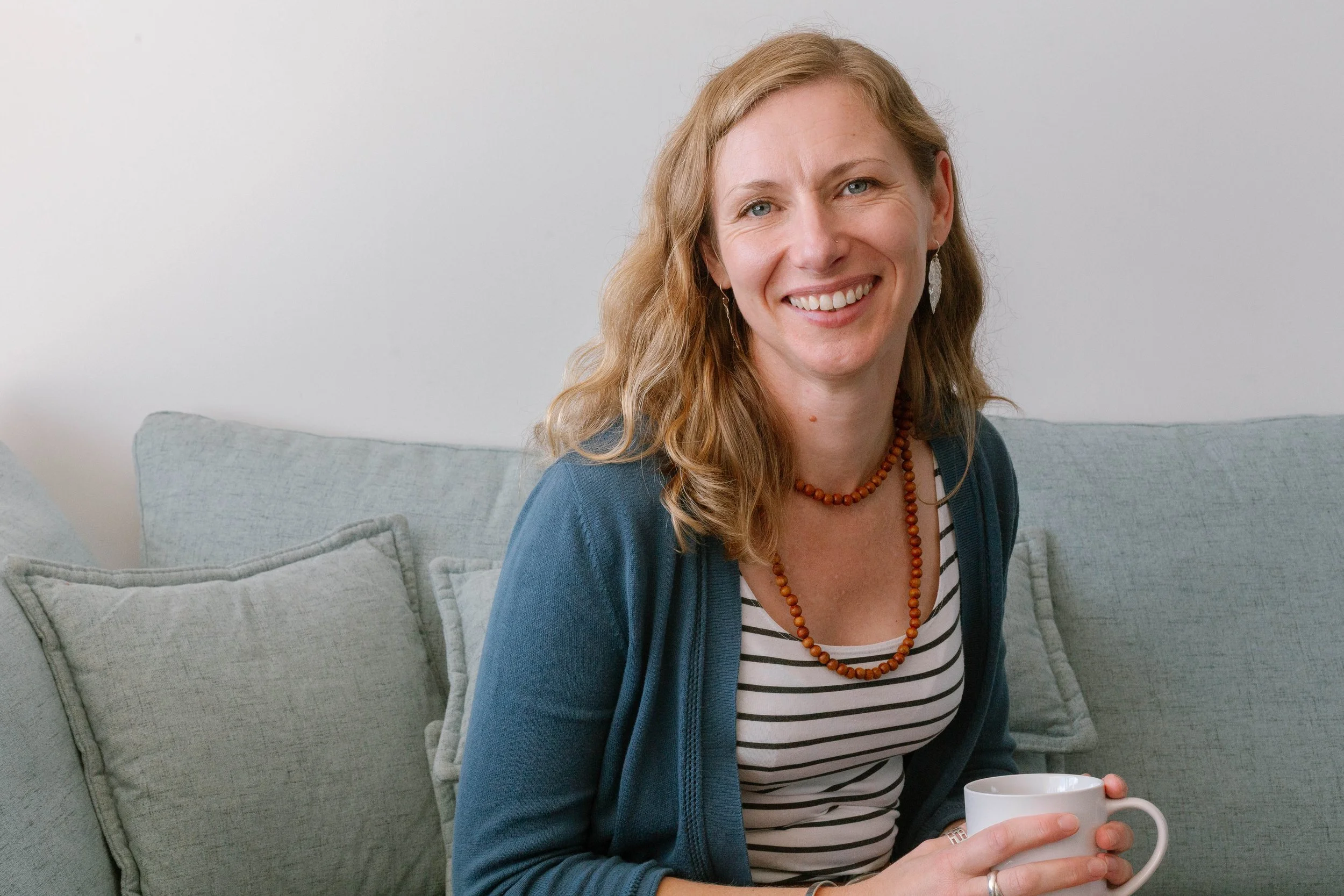 Smiling woman with blonde hair holding a white mug, sitting on gray couch with pillows, against a light-colored wall.