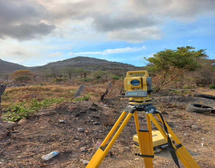 Equipo de topografía amarillo en un paisaje árido con vegetación dispersa y colinas al fondo, cielo parcialmente nublado.