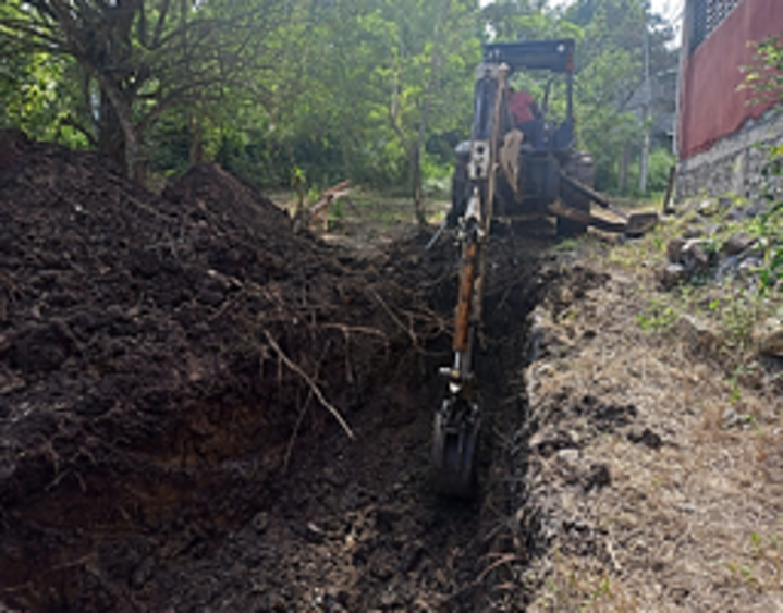 Maquinaria de excavación trabajando en un hoyo en el suelo, con arbustos y árboles en el fondo.