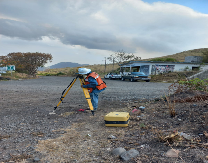 Persona con equipo de topografía usando un teodolito en un área rural con vehículos y edificios en el fondo.