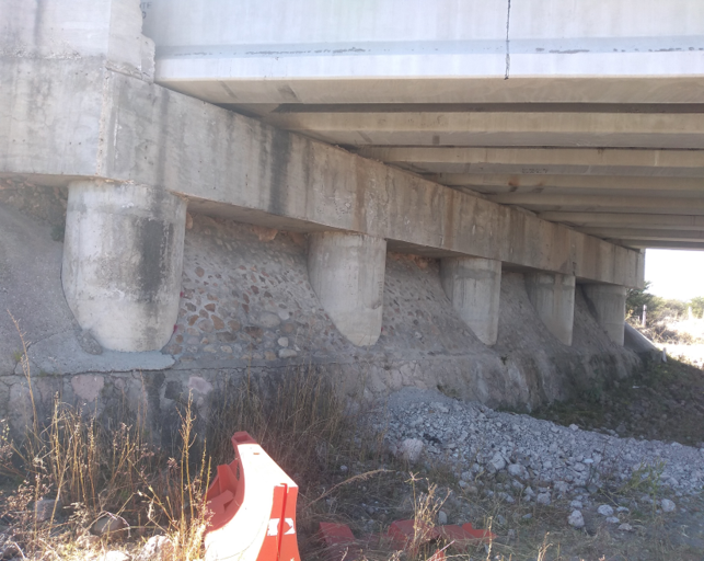 Vista de la estructura de un puente con columnas de concreto y área de tierra y piedras debajo.