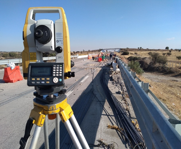 Estación total de topografía en un puente en construcción, con cables y maquinaria en el fondo.