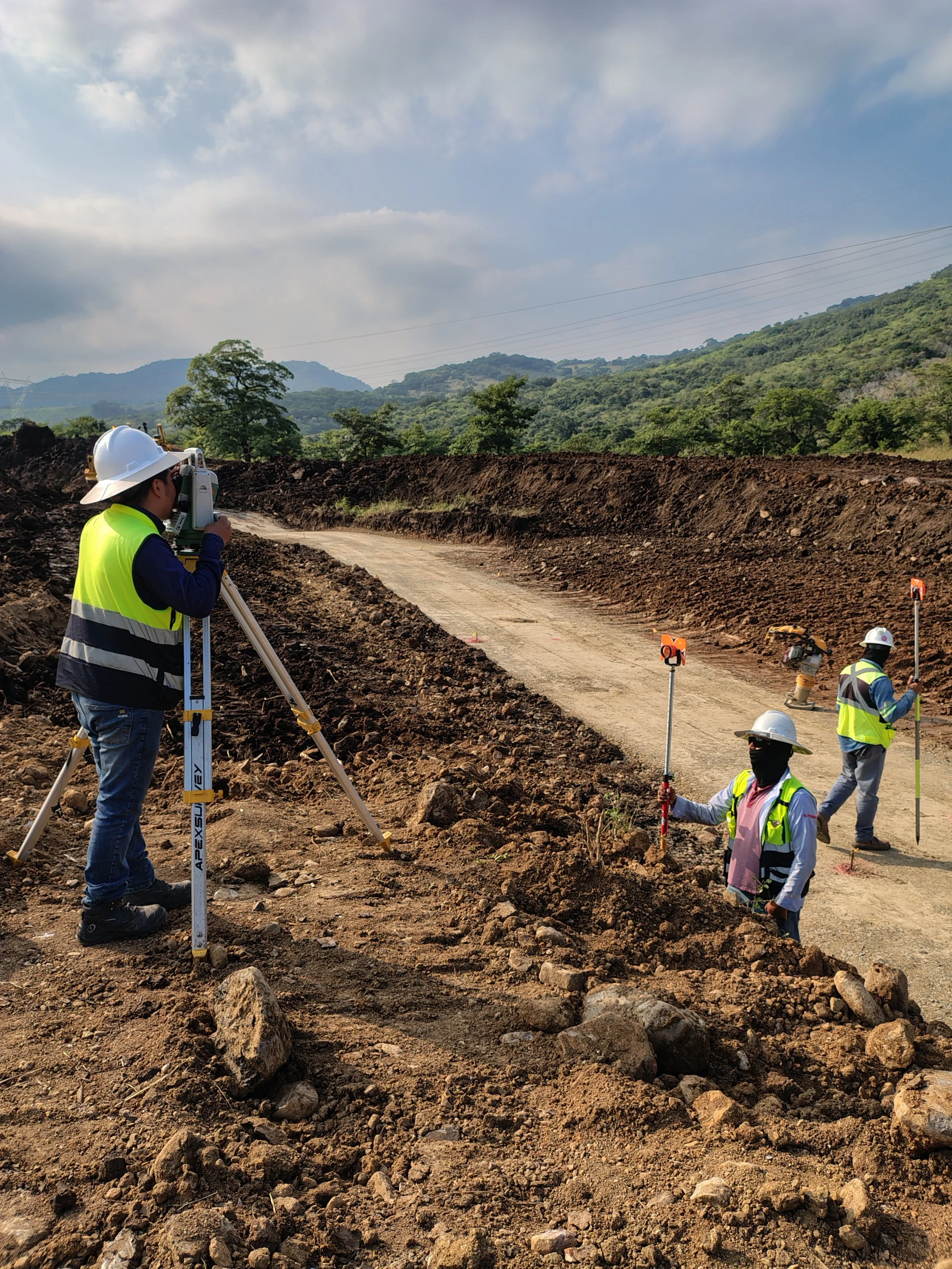 Cuatro trabajadores de construcción en un sitio de obra civil, usando cascos y chalecos reflectantes, uno con nivel de mano, otro con equipo de medición, en un camino en proceso de construcción con fondo de montañas y cielo con nubes.