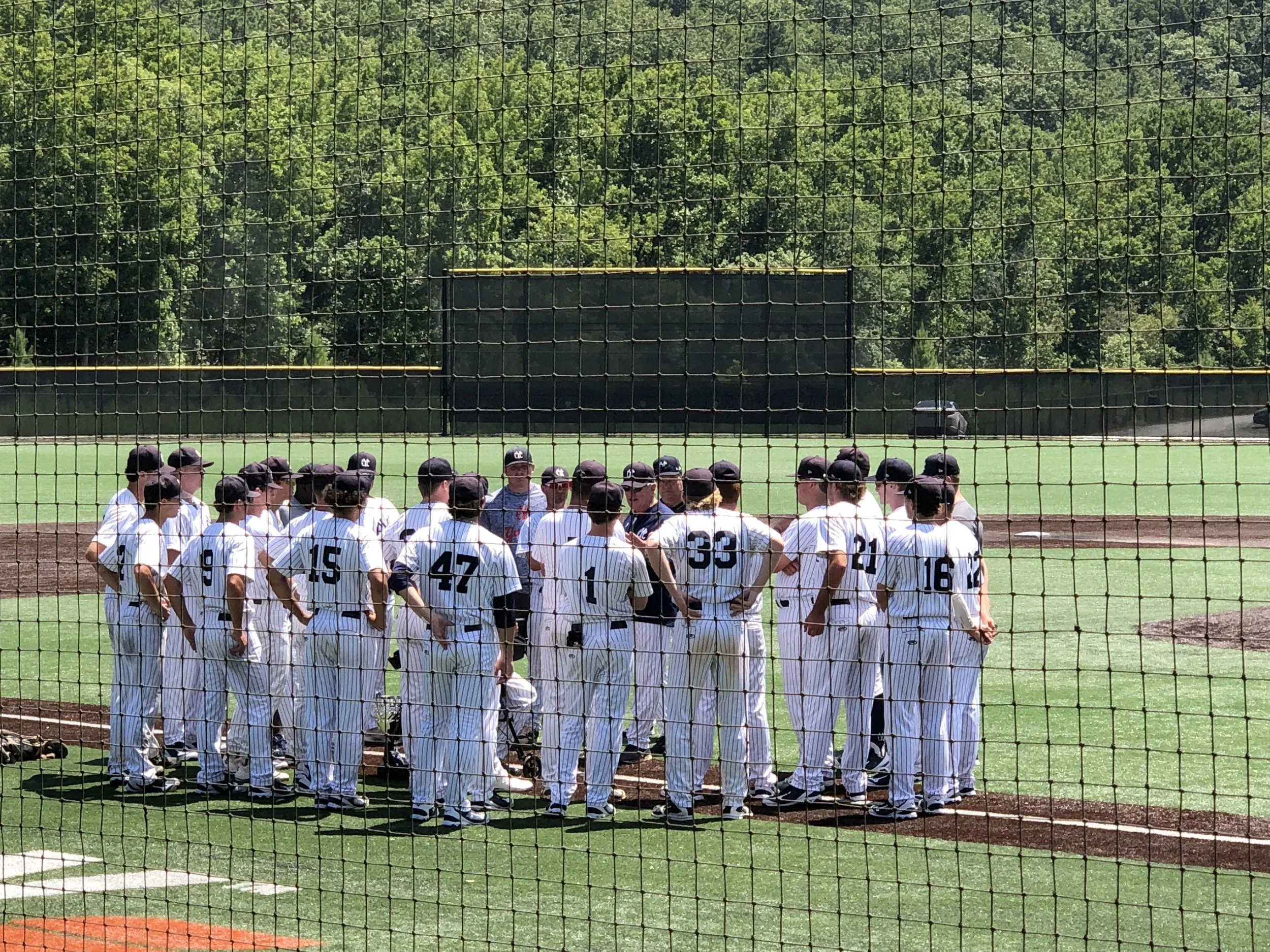 Baseball team huddled on the field during a game, viewed through a protective net.