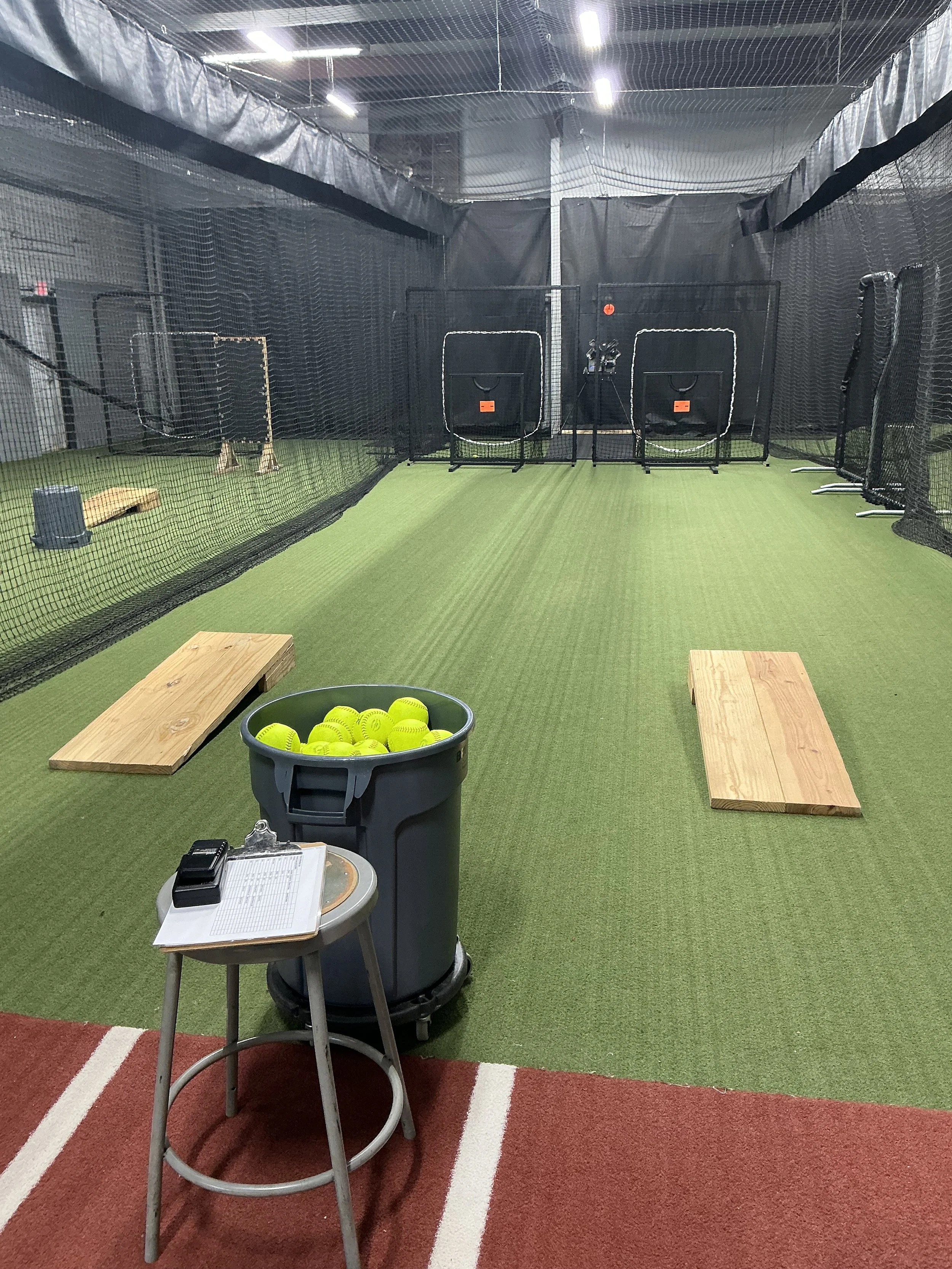 Indoor golf training facility with artificial turf, golf nets, practice targets, and a bucket of tennis balls on a stand in the foreground.