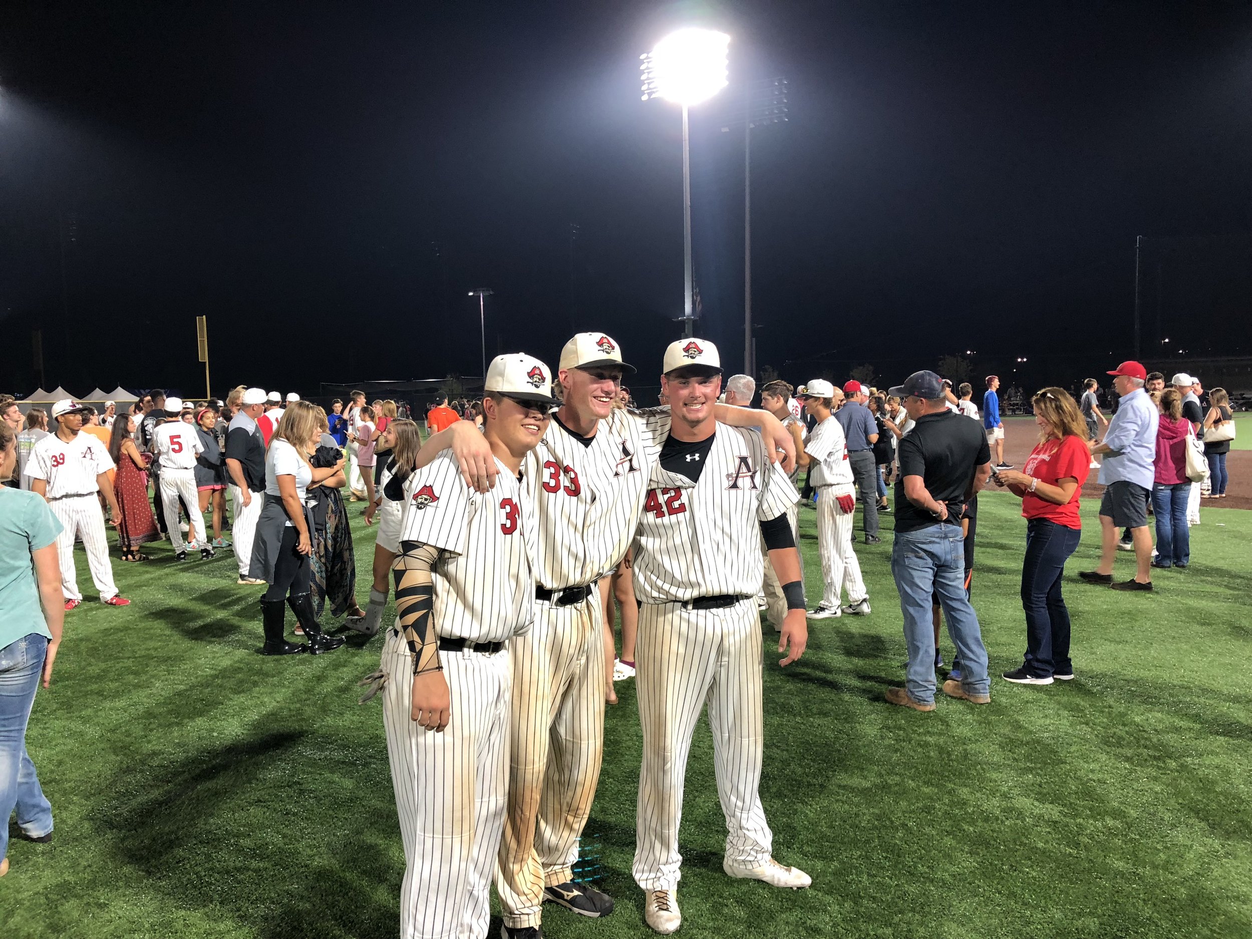 Three baseball players in uniforms with pinstripes and caps standing together on a baseball field at night, crowd in the background.