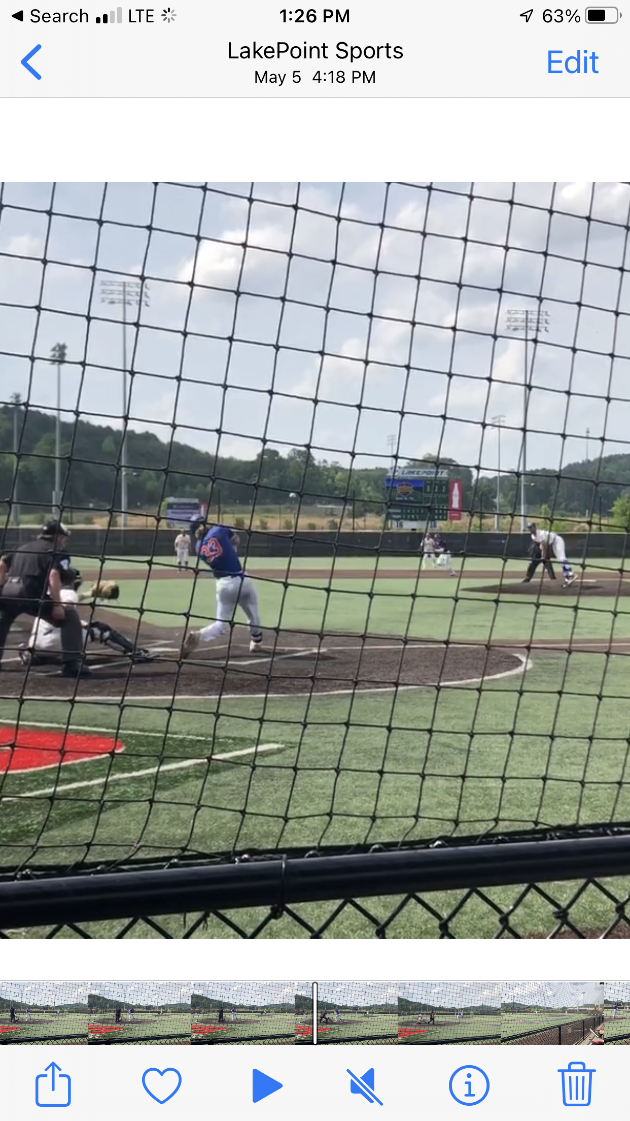 A baseball game mid-play viewed through a protective net, with players on the field, including the batter, pitcher, catcher, and infielders, on a sunny day at LakePoint Sports.