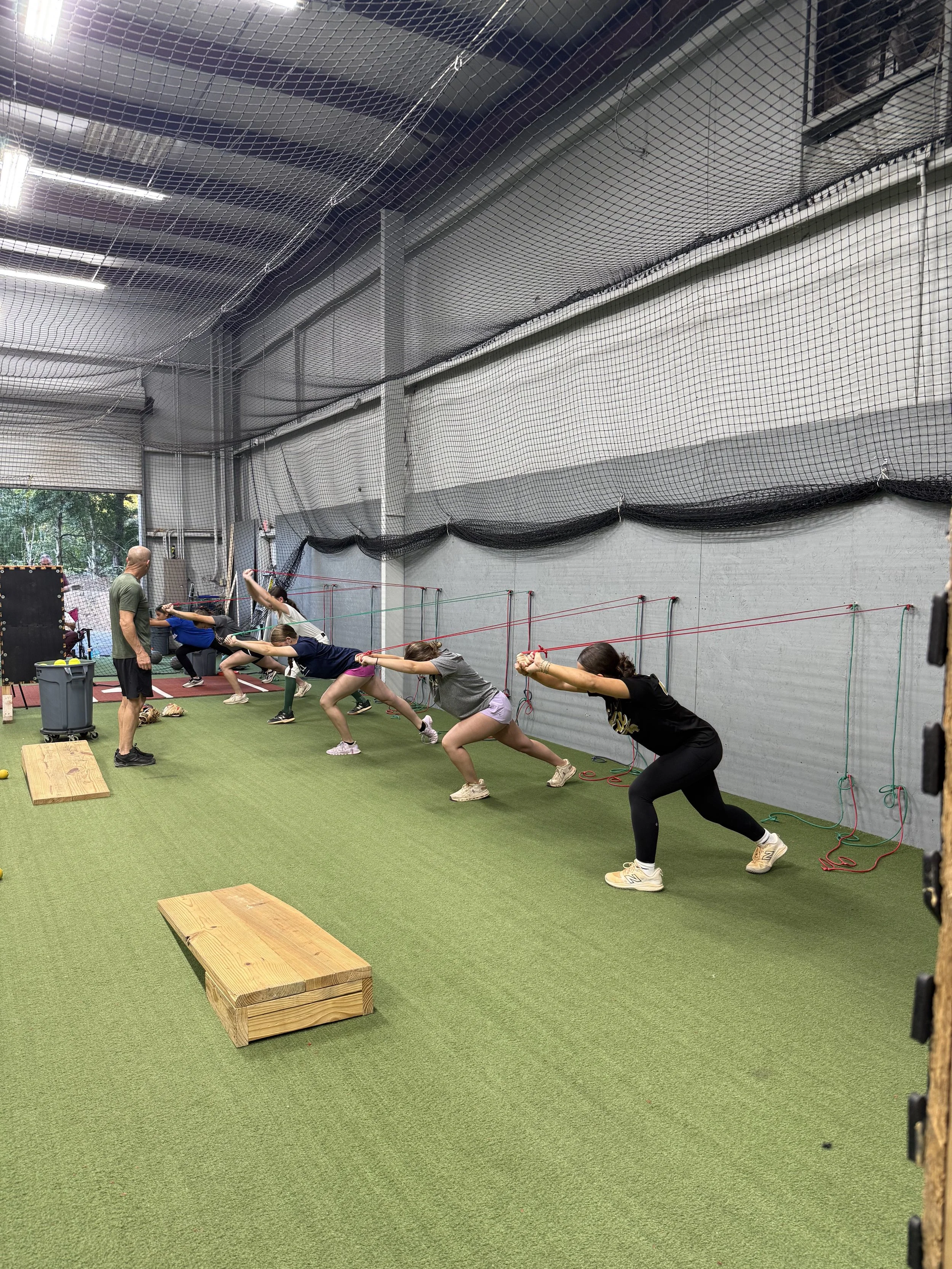 Group of people participating in a resistance training class using resistance bands in an indoor gym with artificial turf flooring and a coach supervising.