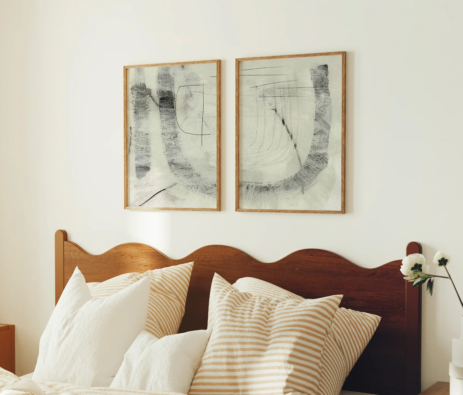 A bedroom with two abstract black and white paintings above a wooden bed frame. The bed has striped and white pillows, and a vase with white flowers is on a nightstand next to the bed.