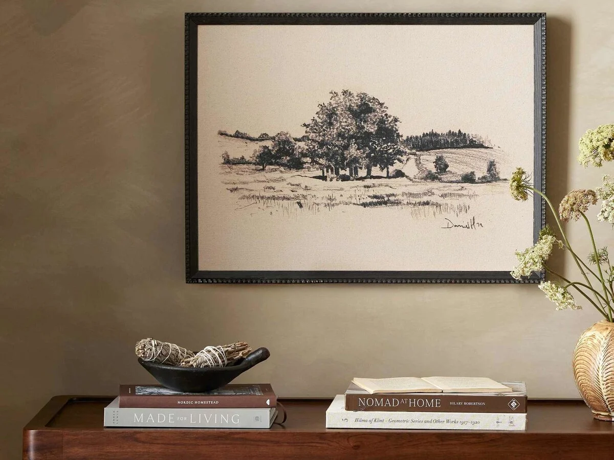 A framed black and white landscape sketch of trees and fields on a beige wall. Below, on a wooden table, there is a black bowl with bundles of twine or dried herbs, and a stack of books including titles like "Nordic Homestead," "Made for Living," and