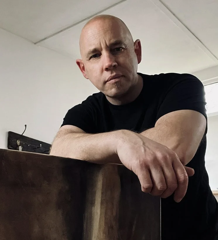 A bald man in a black t-shirt leaning on a wooden surface, looking directly at the camera with a serious expression, in an indoor setting.