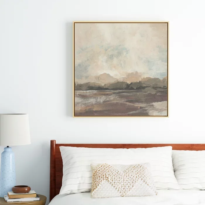 A bedroom with a framed landscape painting above the bed, consisting of neutral tones. There is a white pillow with a textured, patterned throw pillow on a wooden headboard. To the left is a small bedside table with a blue lamp and some books.