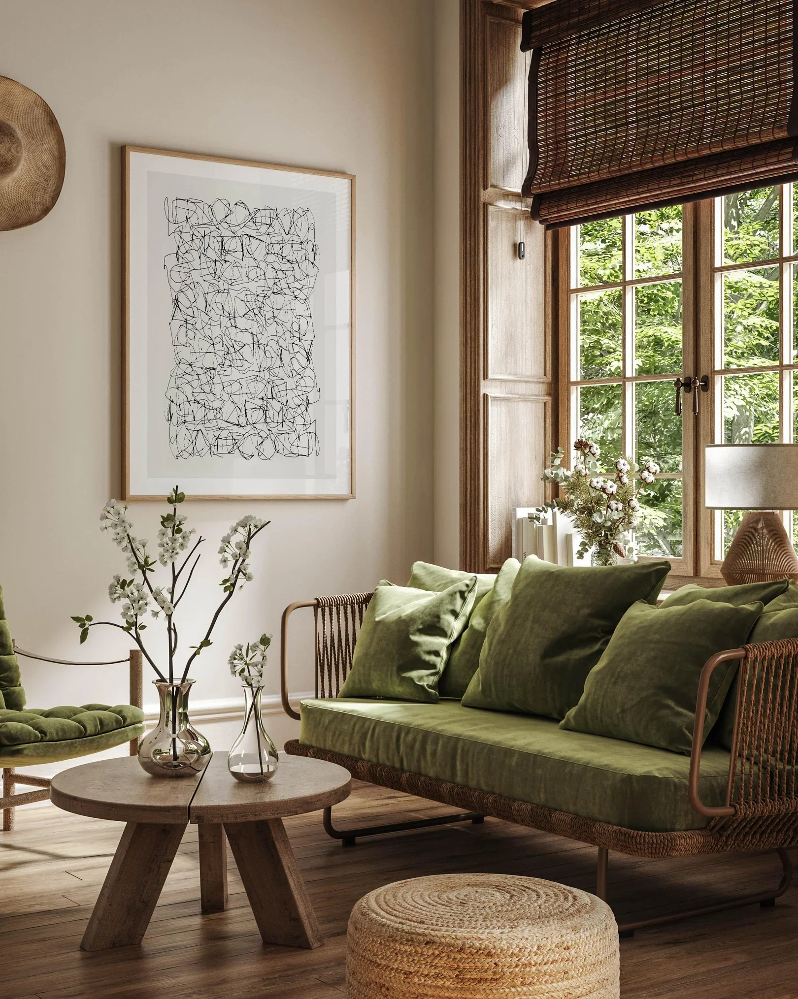 A living room with a green sofa, matching pillows, a wooden round coffee table, and an armchair. There are vases with white flowers on the table, a large framed abstract black and white artwork on the wall, and a window with wooden trim and blinds showing green outside scenery.
