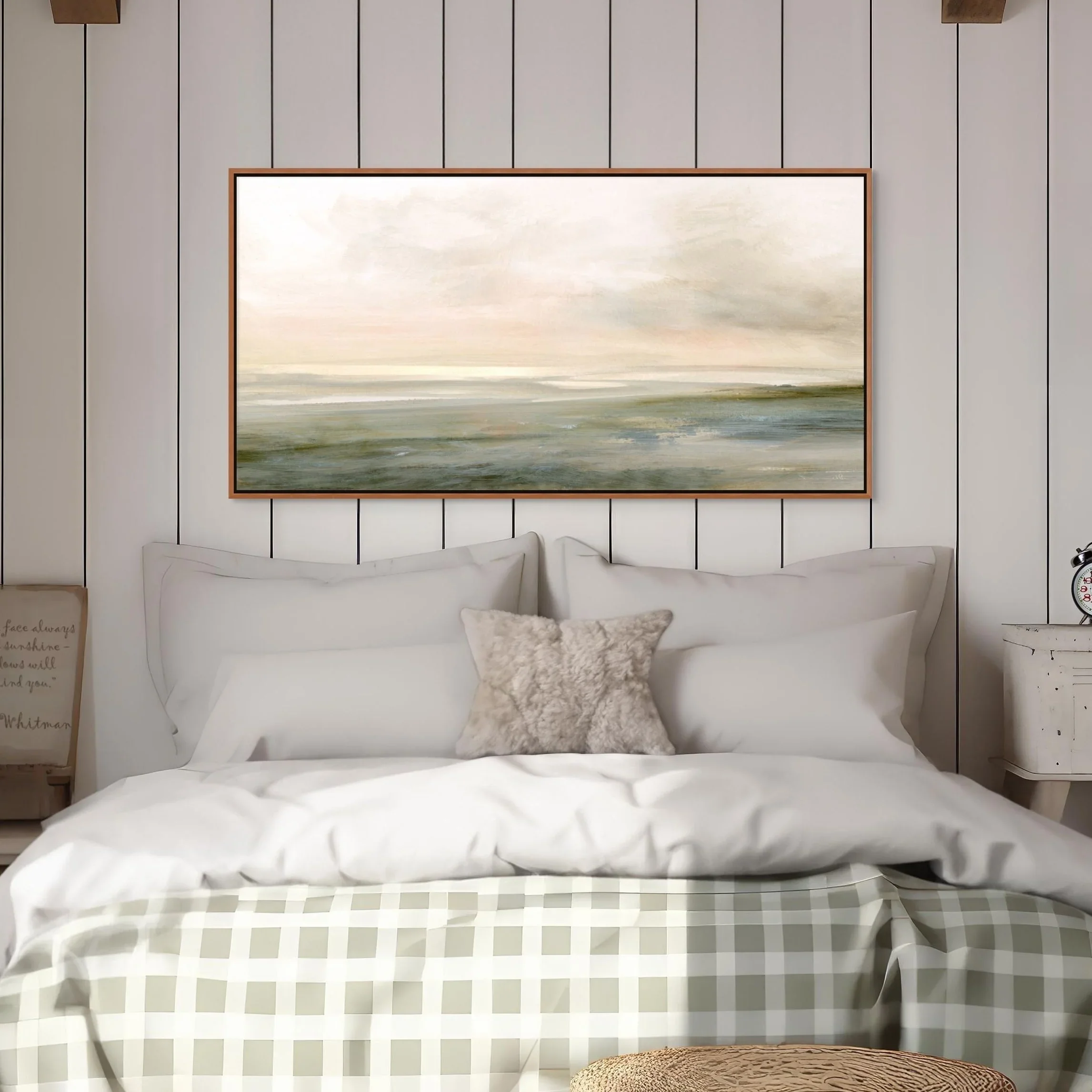A cozy bedroom with a bed, white pillows, a furry gray decorative pillow, and a green checkered bedspread. Above the bed is a large, framed landscape painting with soft, muted colors. On the left, a small wooden sign leans against the wall, and on th