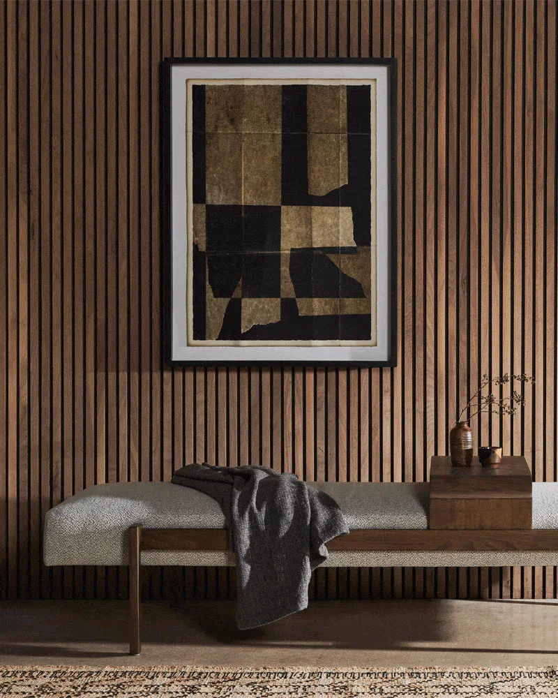 Modern interior with a wooden paneled wall, a framed abstract art piece, a bench with a gray cloth draped over it, and a wooden table with decorative vases and dried branches.