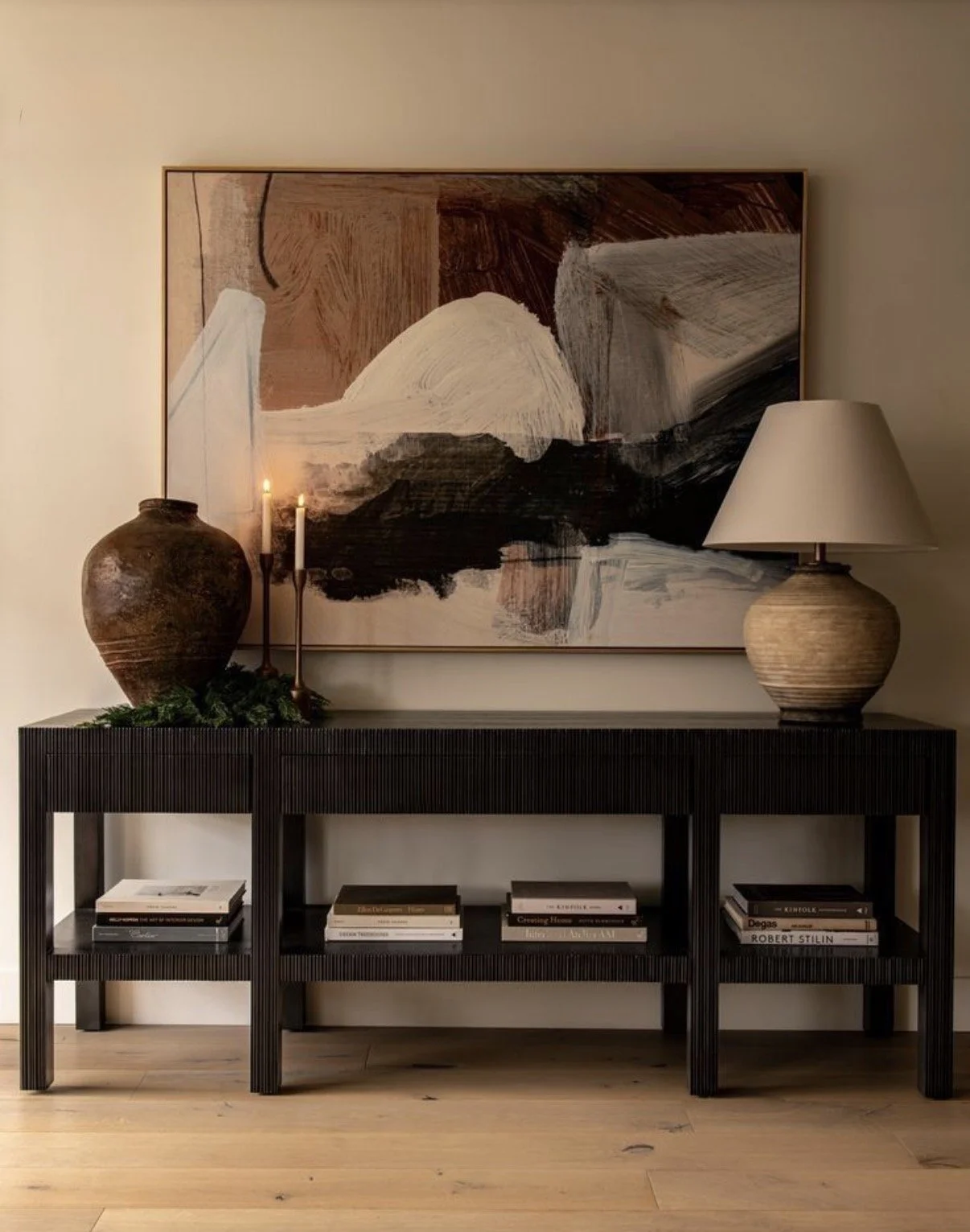 A black console table with a textured surface, decorated with two large pottery vases, two lit candles, and a small green plant. Above the table hangs an abstract painting with earth tones, and to the right, a large beige ceramic table lamp.
