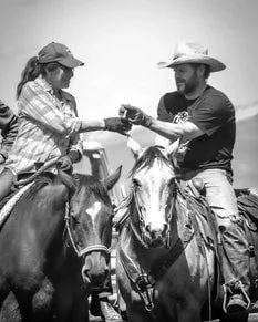 A man and a woman on horseback exchanging a drink during a cattle ranching activity, both wearing cowboy hats.