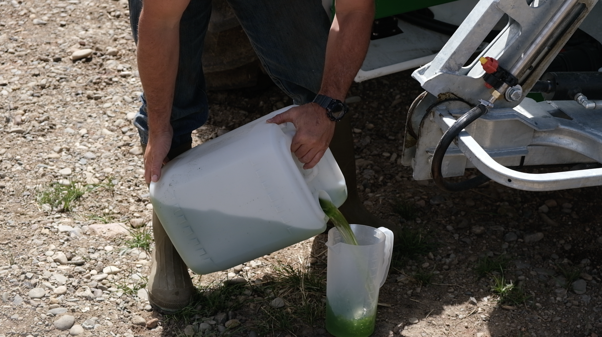 A person is pouring a green liquid from a white plastic container into a smaller clear plastic container, outdoors on rocky ground.