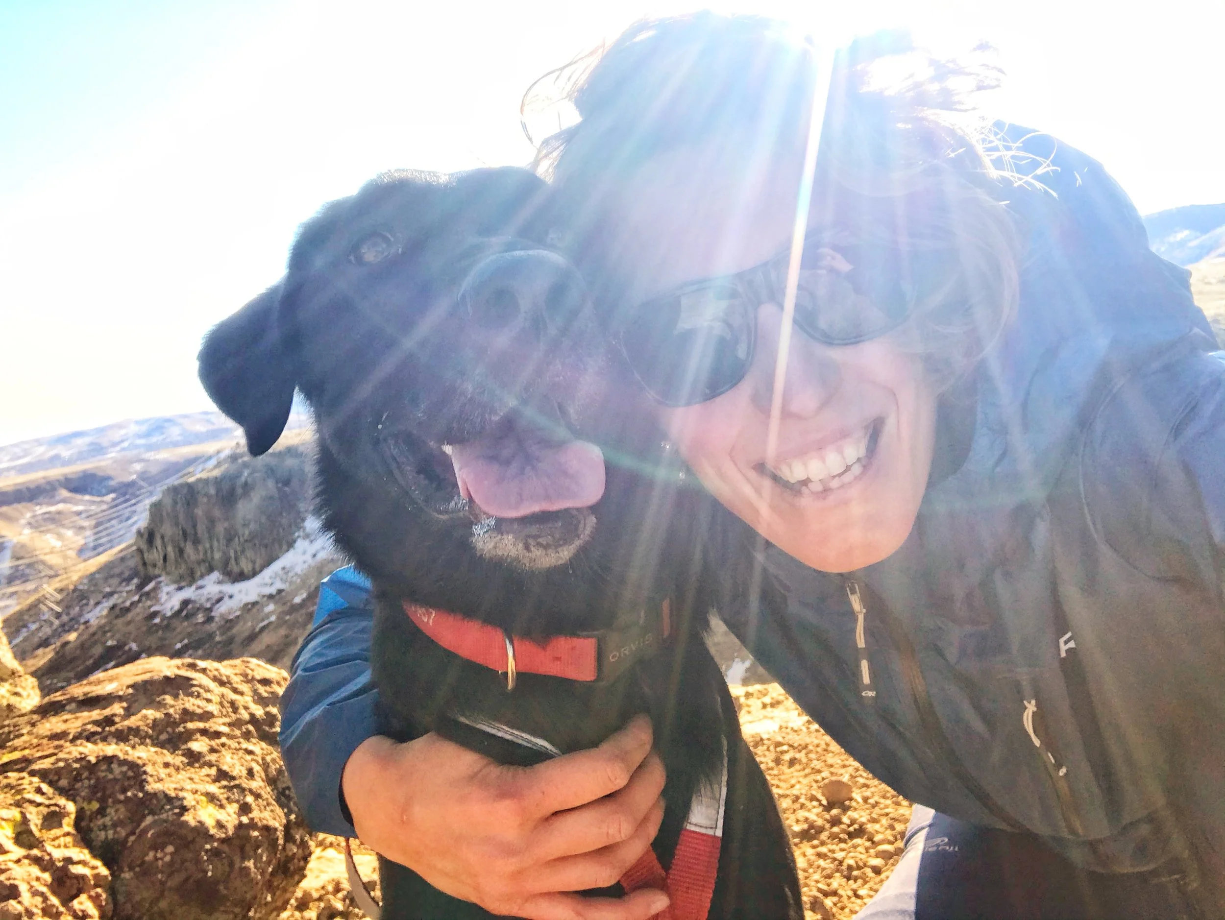 A woman wearing sunglasses and a jacket with a mountain landscape in the background, smiling while hugging a black dog with a red collar.