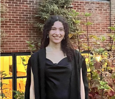 A young woman smiling outdoors in front of a brick wall and plants, wearing a black dress and graduation gown.