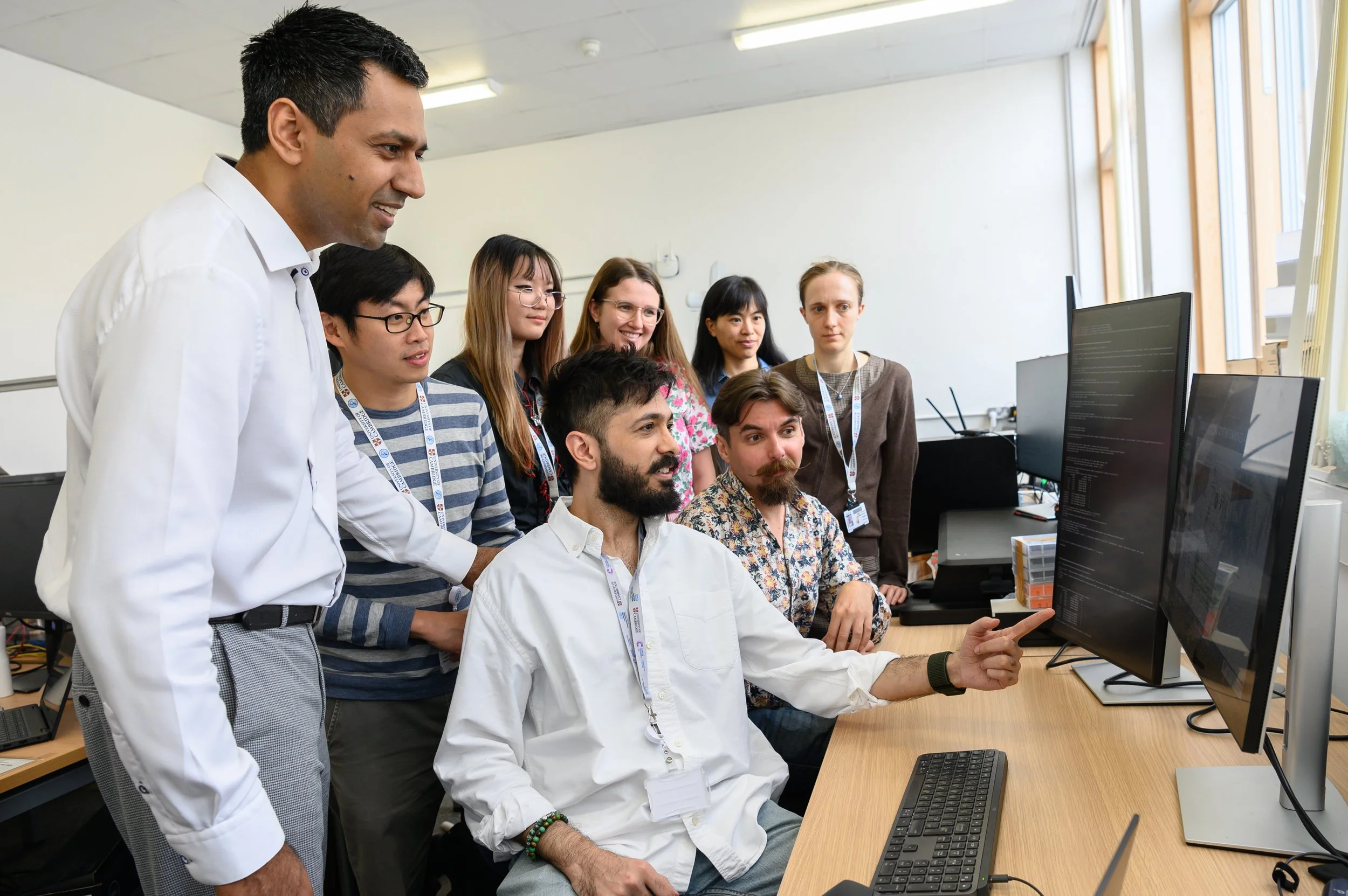 A group of diverse young professionals and colleagues gathered around a seated man pointing at a computer screen in an office, with multiple monitors and a bright window in the background.
