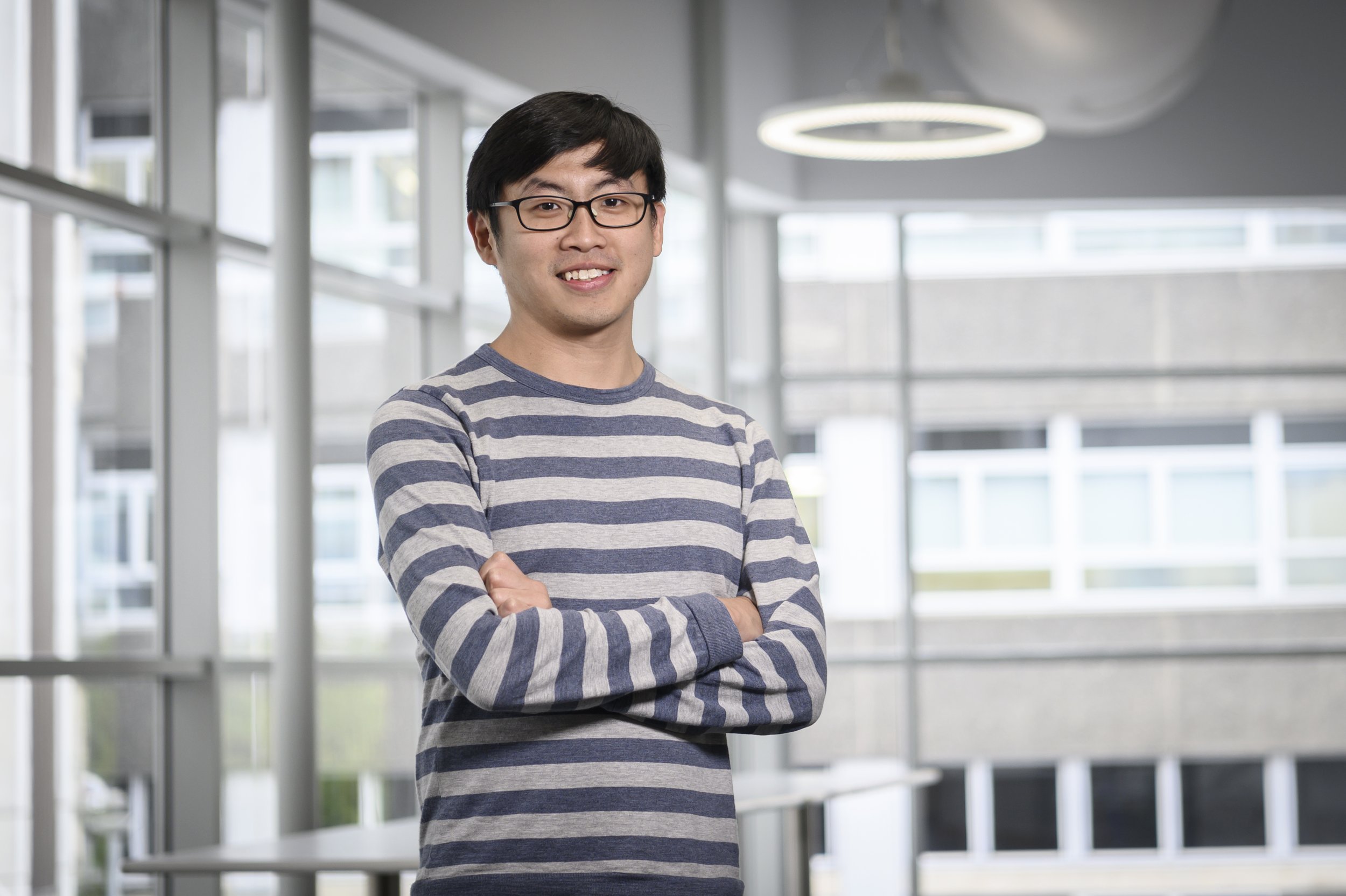 A young man with glasses and black hair smiling, standing with arms crossed in a modern indoor space with large windows and architectural lighting.