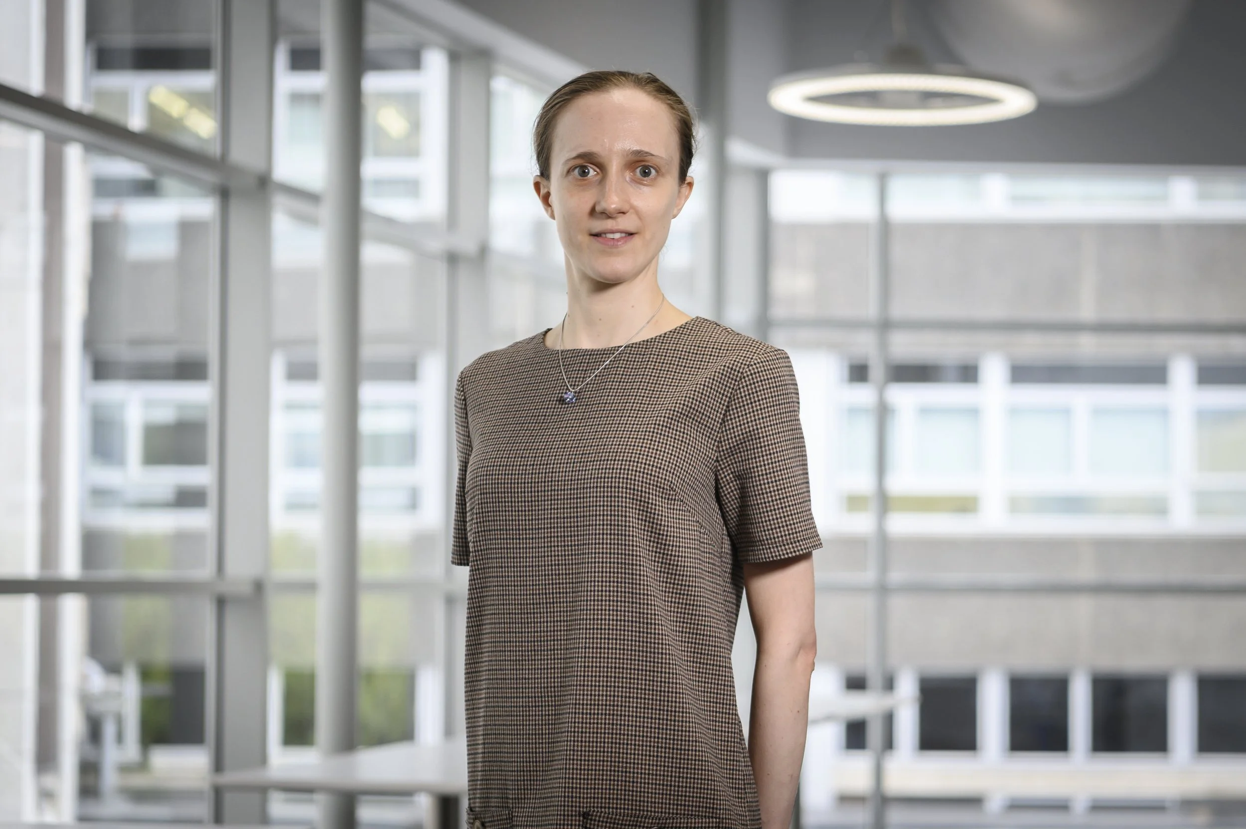 A woman with brown hair in a ponytail wearing a brown checkered dress standing indoors with a modern glass building in the background.