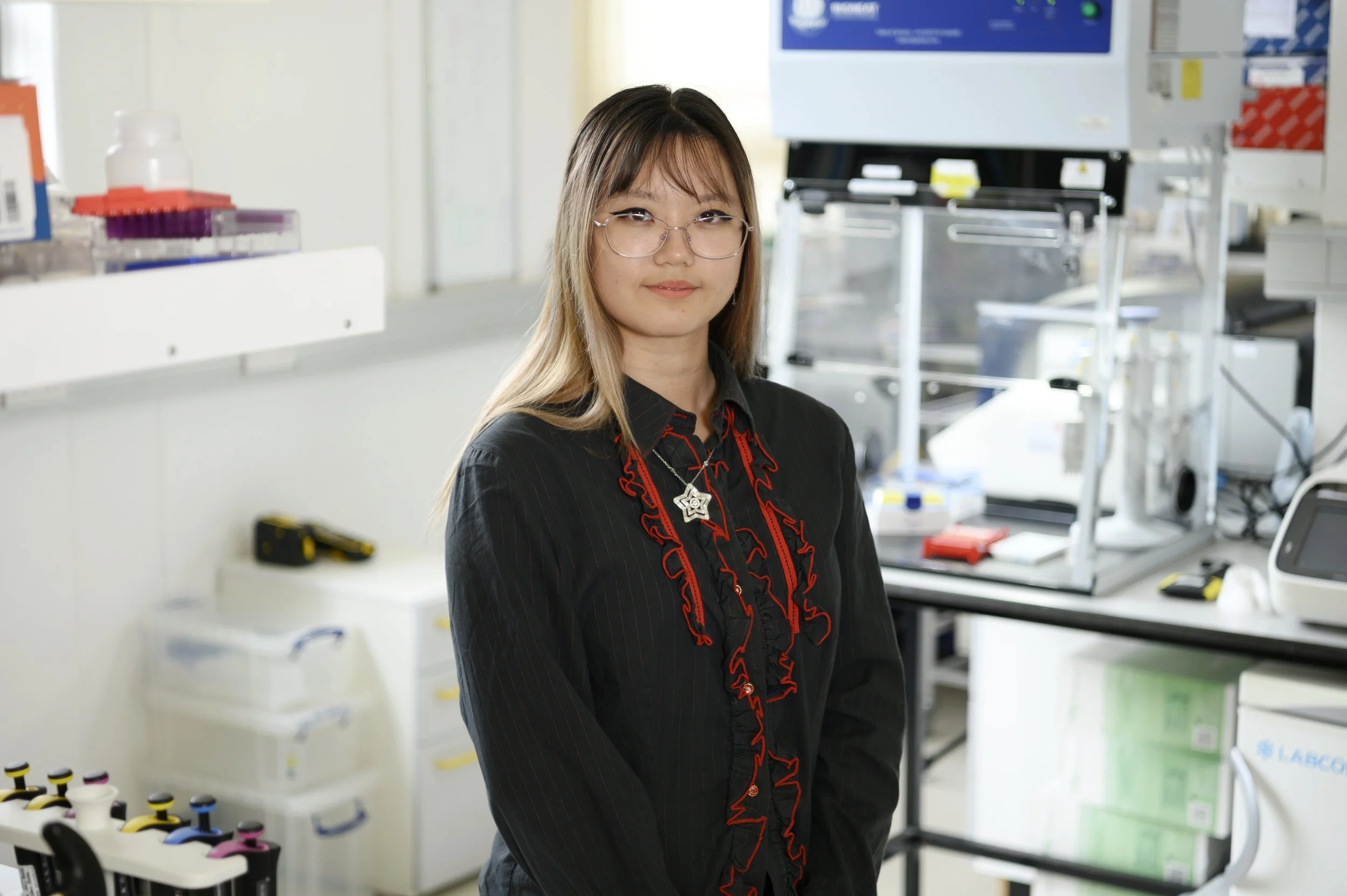 A young woman with glasses and straight hair stands indoors in front of large windows and modern lighting, wearing a dark pinstripe shirt with ruffles.