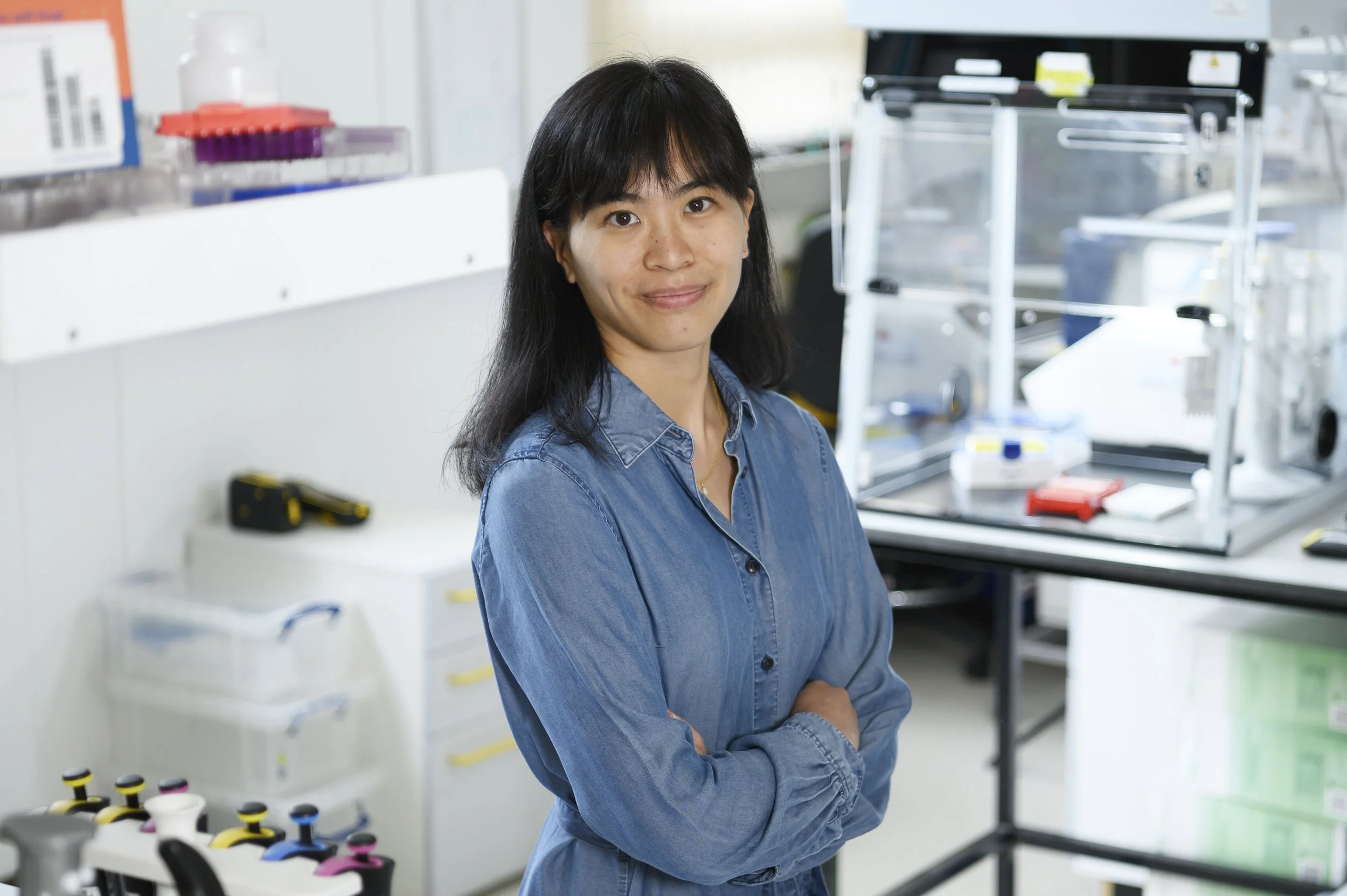 A black-and-white photo of a woman with shoulder-length hair wearing a button-down dress, standing indoors near large windows with architectural details in the background.