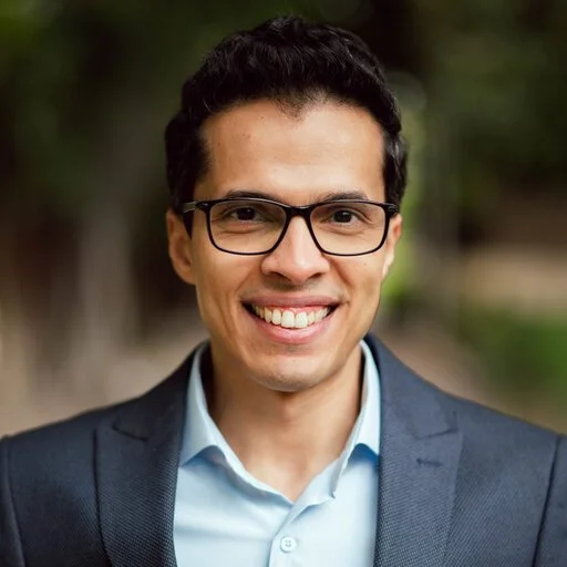 A man with dark hair, glasses, and a bright smile wearing a navy suit jacket and light blue dress shirt outdoors with trees in the background.