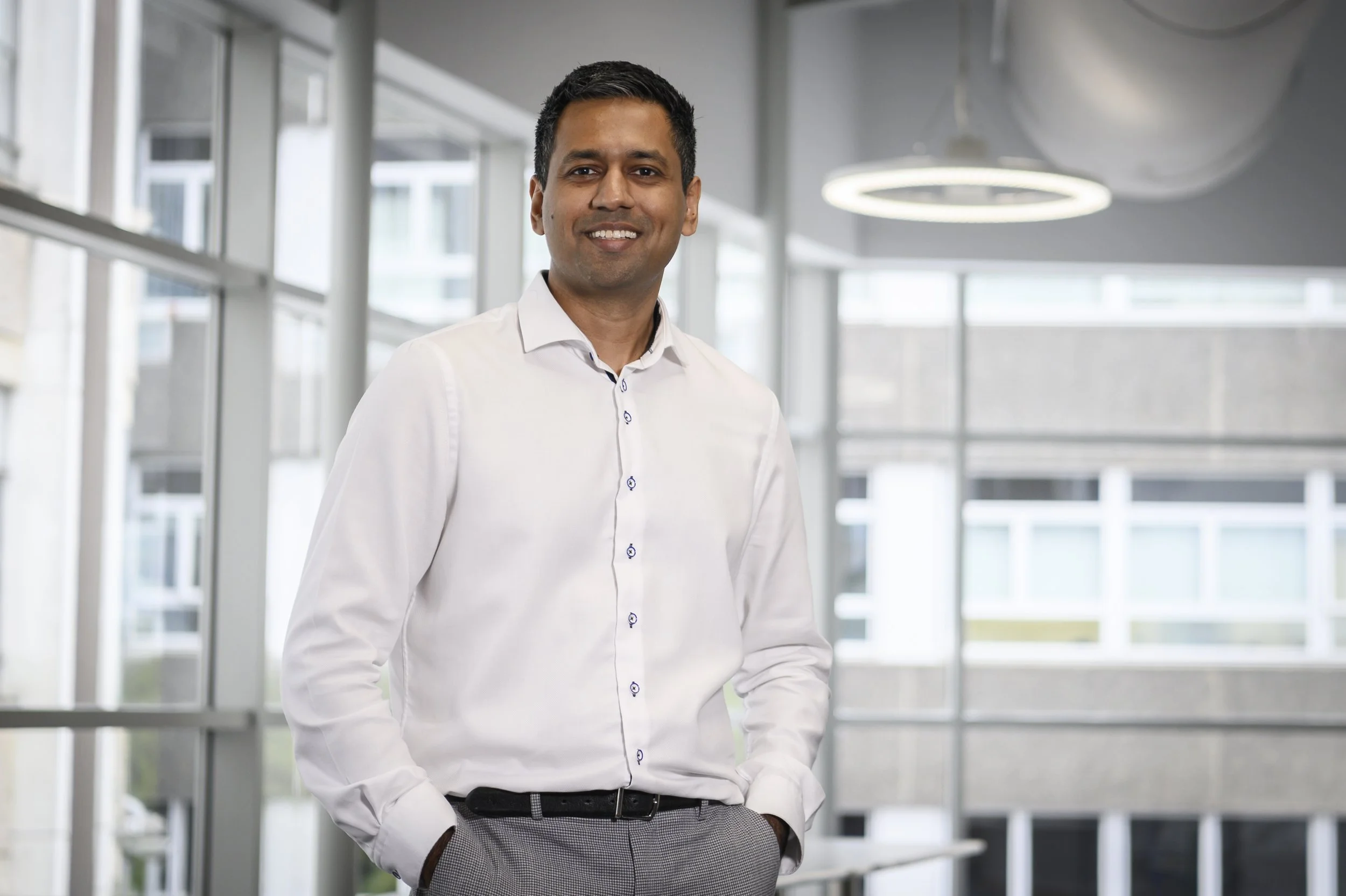 A smiling man in a white dress shirt and gray pants stands in a modern office with large windows and circular ceiling lights.
