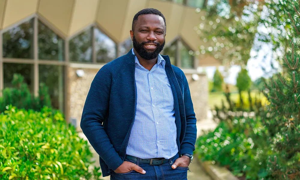 A man with a beard smiling outdoors, wearing a casual jacket over a button-up shirt, with hands in his pockets, standing on a sidewalk with trees and a modern building with glass windows in the background.
