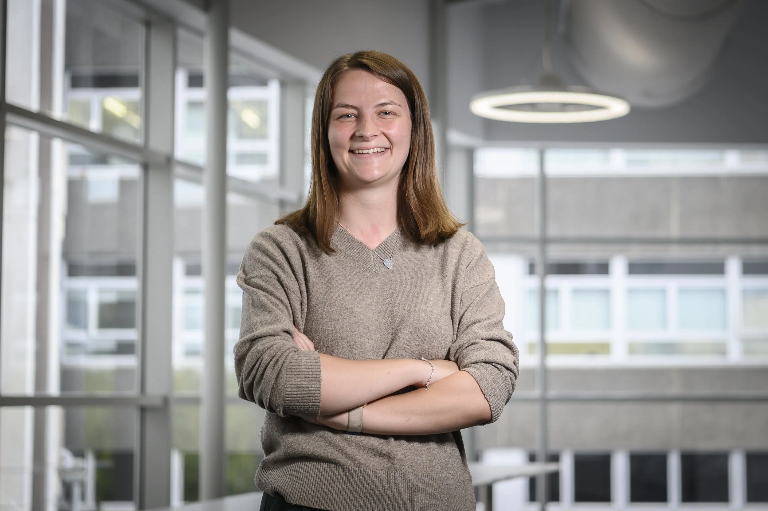 A smiling young woman with brown hair wearing a beige sweater, standing with arms crossed in a modern, well-lit office or university building with large windows.