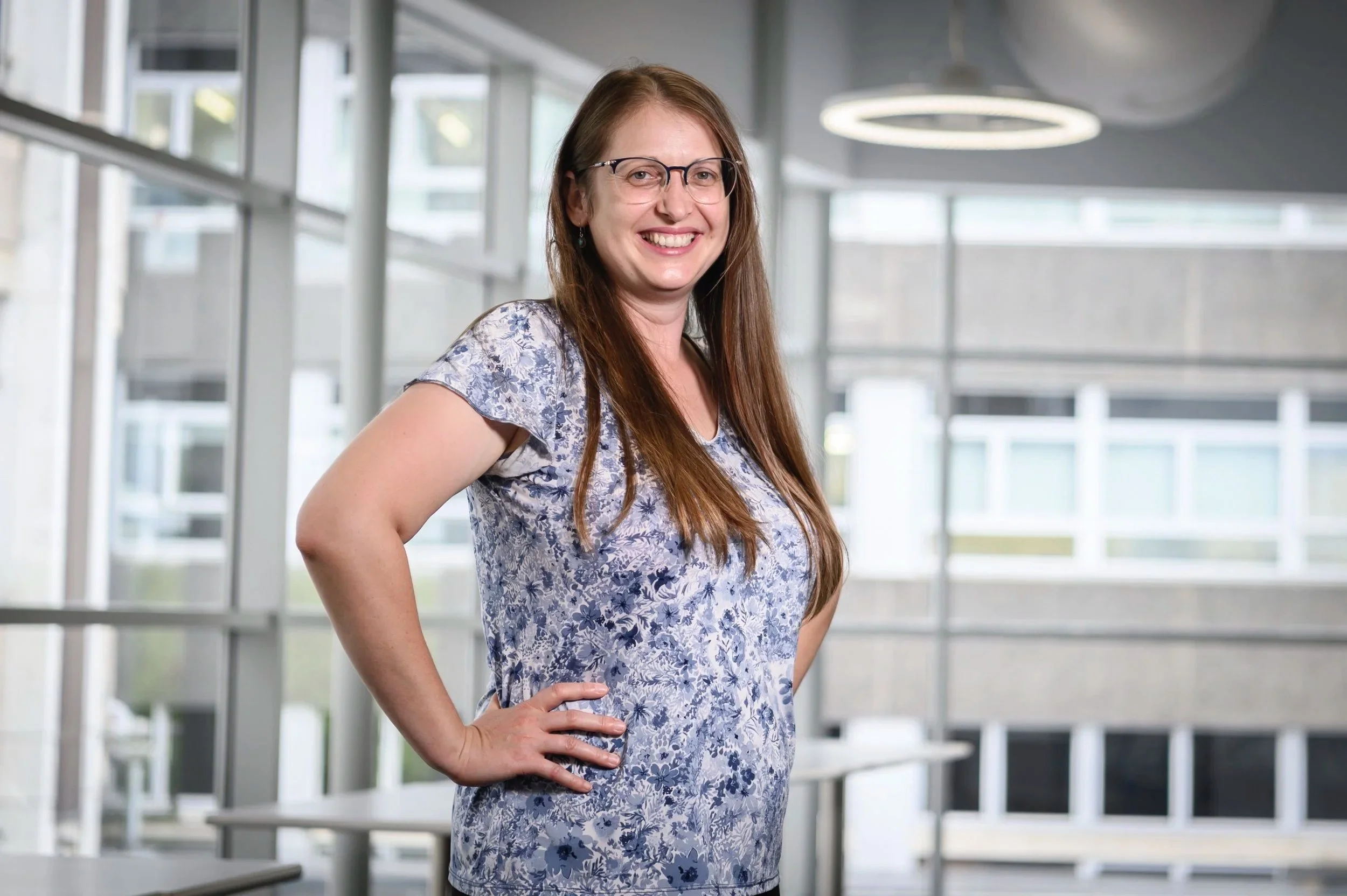 A woman with long brown hair, glasses, and a floral shirt standing indoors with her hand on her hip, smiling at the camera.