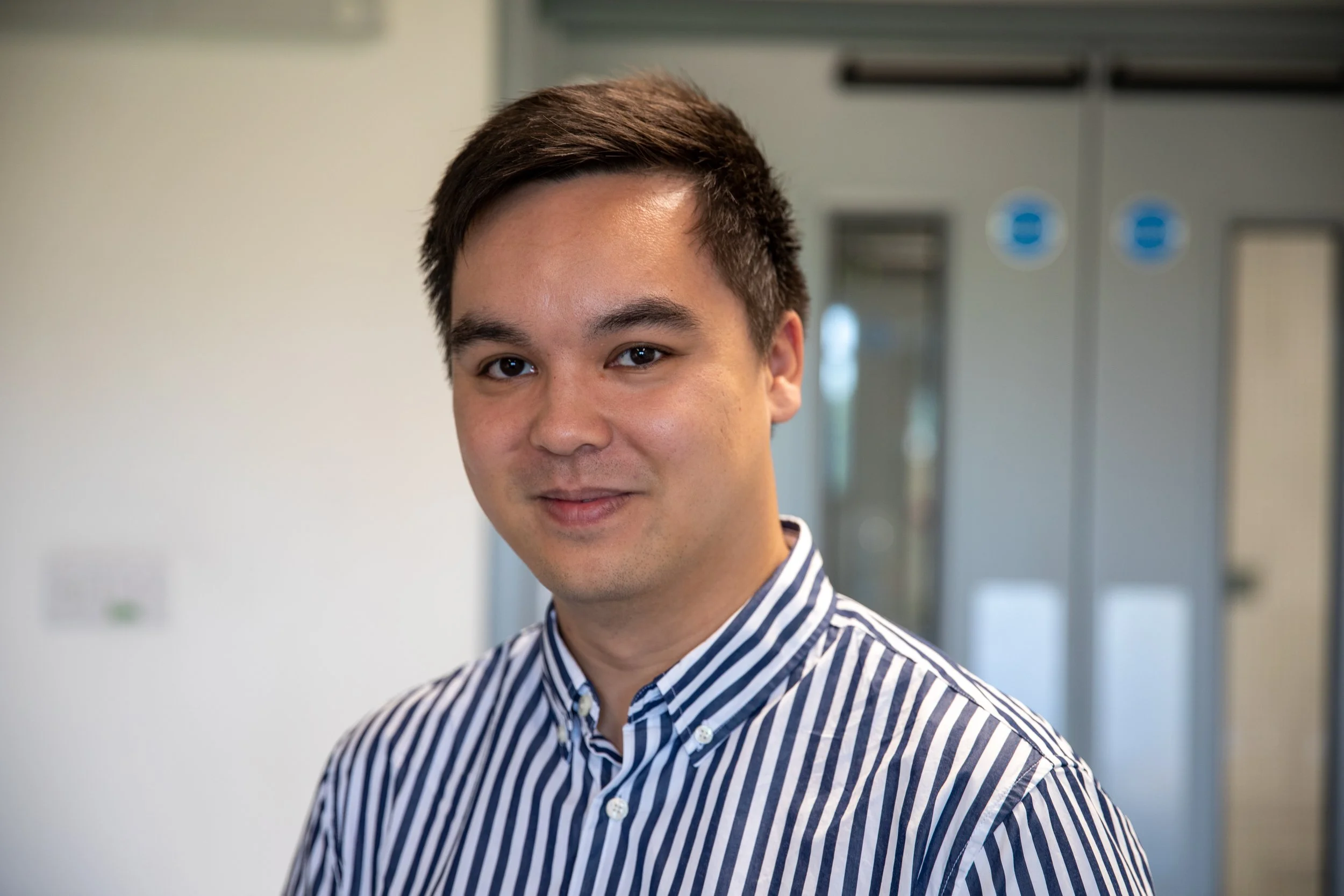 Black and white photograph of a young man wearing a striped shirt, smiling softly, standing indoors in front of a blurred background with office or institutional doors.