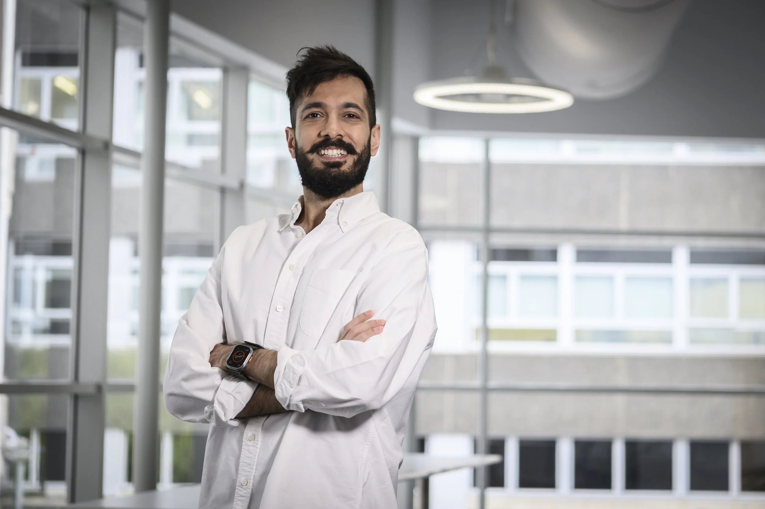 A man with a beard and smart watch standing with arms crossed inside a modern building with large windows.