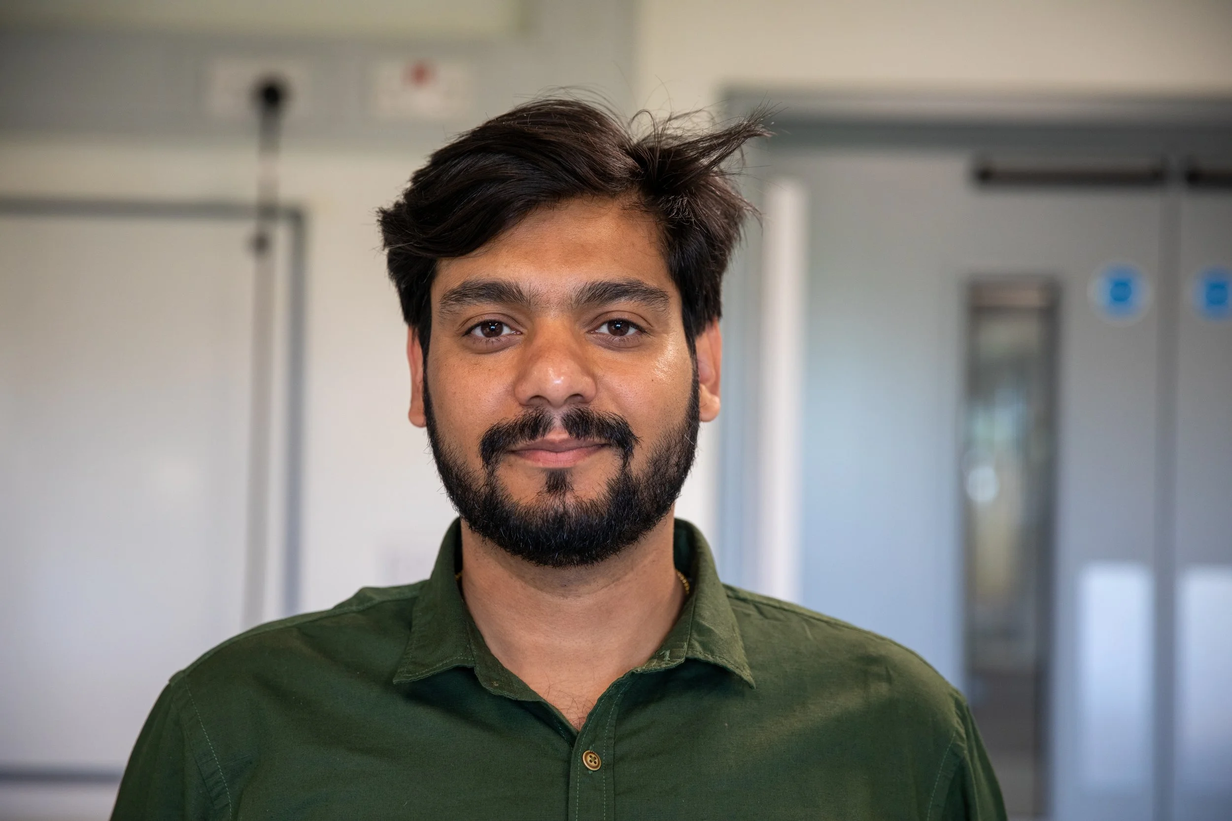 Black and white portrait of a man with dark, slightly messy hair and a beard, wearing a dark collared shirt, standing indoors with a blurred background.