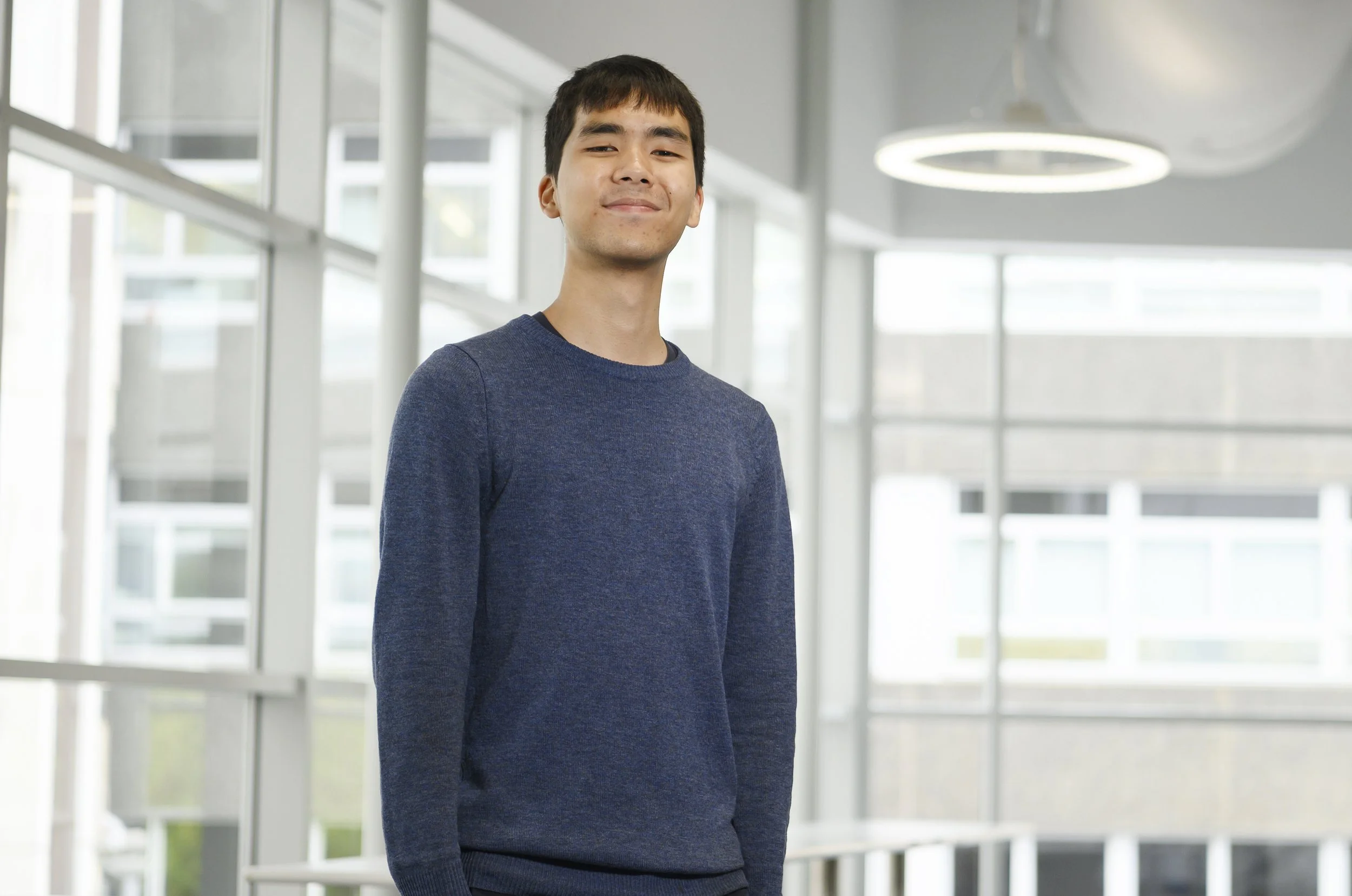 Young man in a blue sweater smiling inside a modern, glass-walled building.