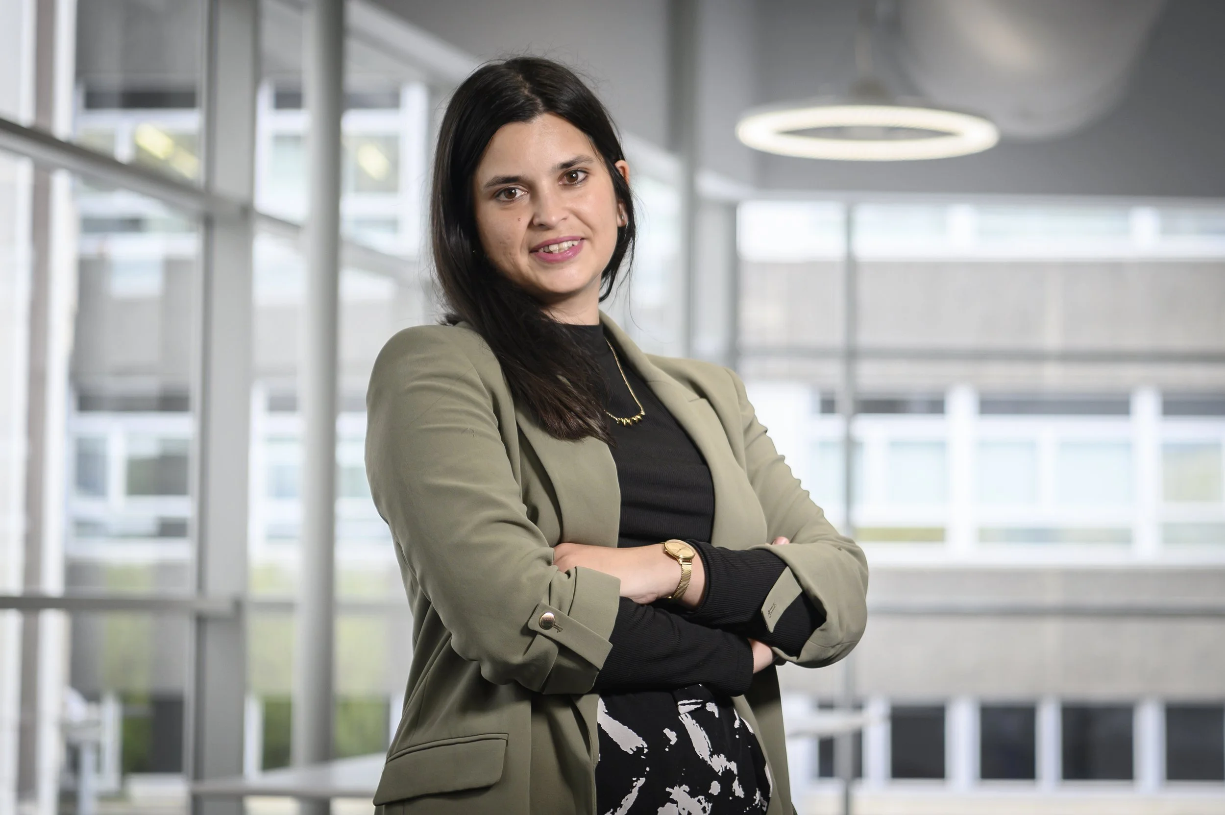 A woman with black hair, wearing a green blazer, black top, and patterned pants, standing with her arms crossed in a modern office building.