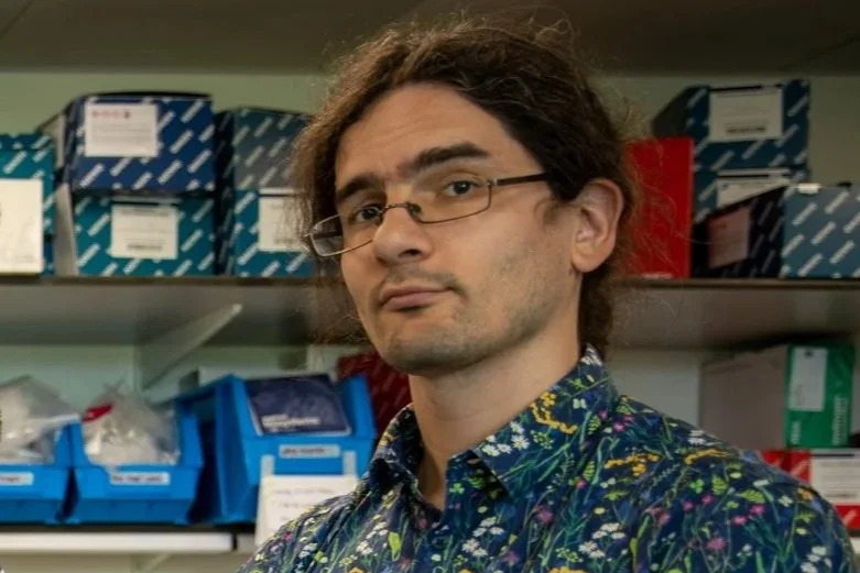 A man with long hair, glasses, and a patterned shirt standing in a room with shelves filled with boxes and supplies.
