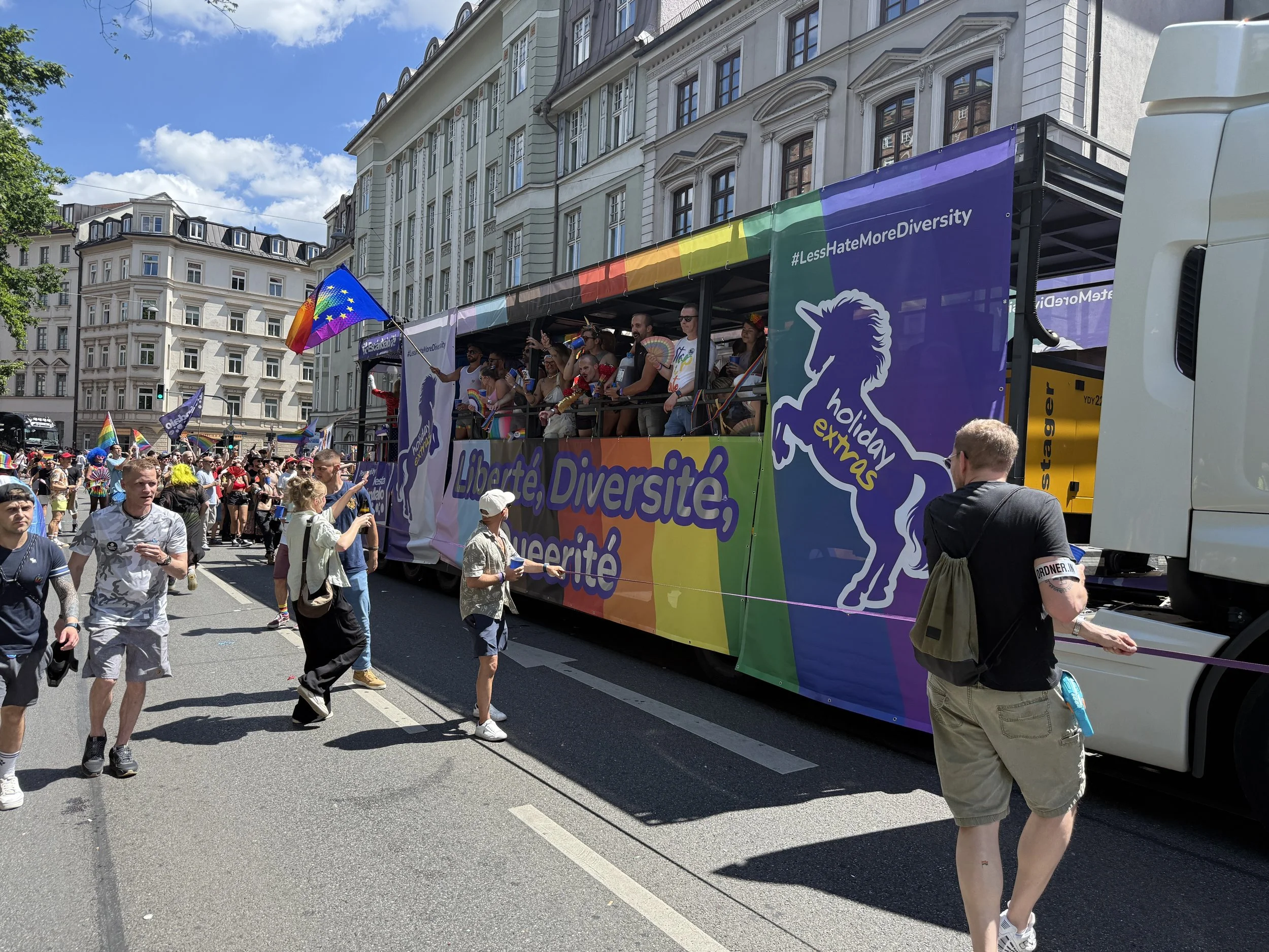 Bunt geschmückter Wagen bei der Pride-Parade in einer Stadt mit Menschen, die aus dem Wagen und vom Straßenrand zuschauen, viele tragen Regenbogen-Fahnen und -Accessoires. Das Banner auf dem Wagen trägt die Wörter 'Liberté, Diversité, Superité' und das Hashtag '#LessHateMoreDiversity'.