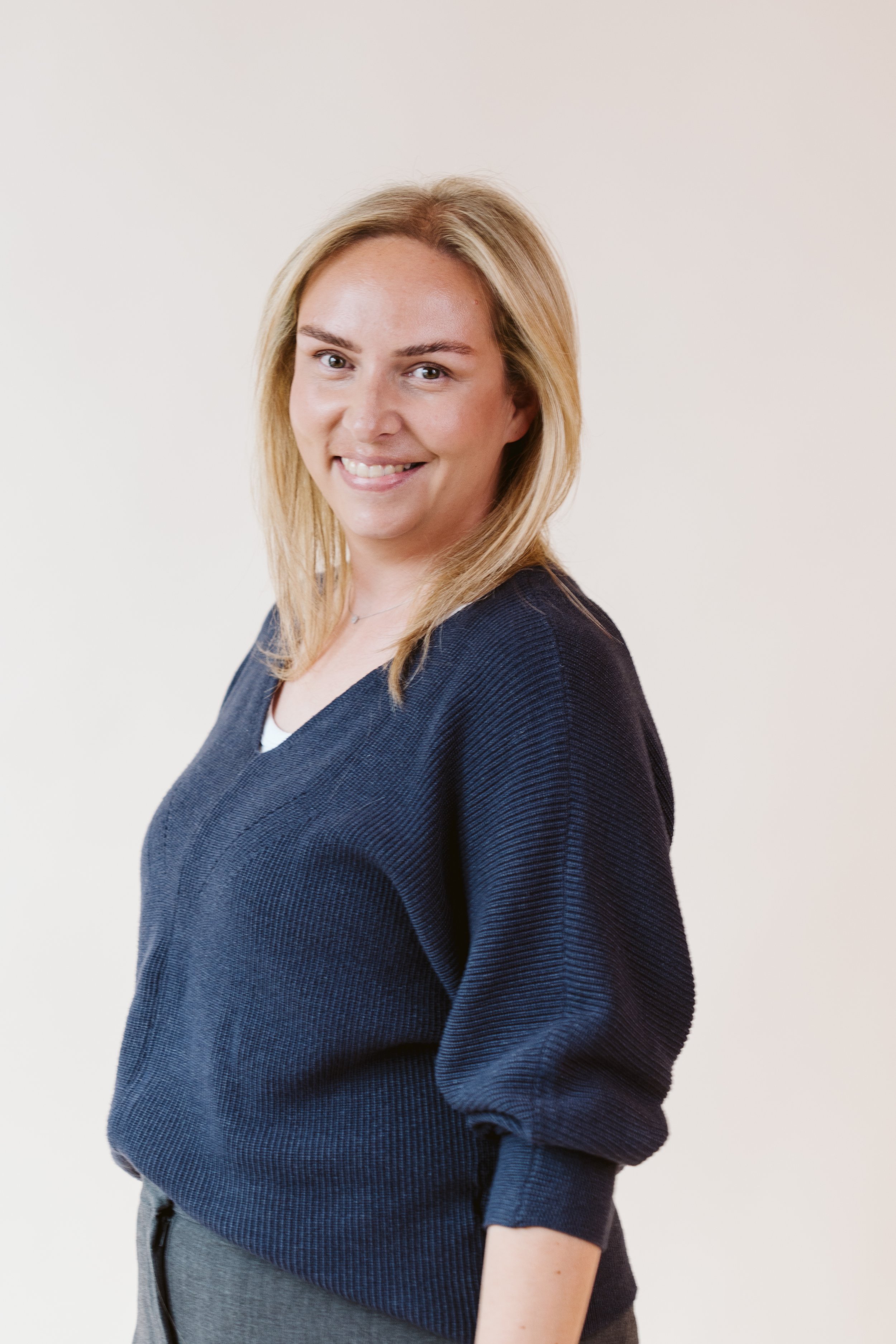 Junge Frau mit blonden Haaren, lächelnd, trägt einen dunkelblauen Pullover und eine weiße Bluse, vor hellem Hintergrund.